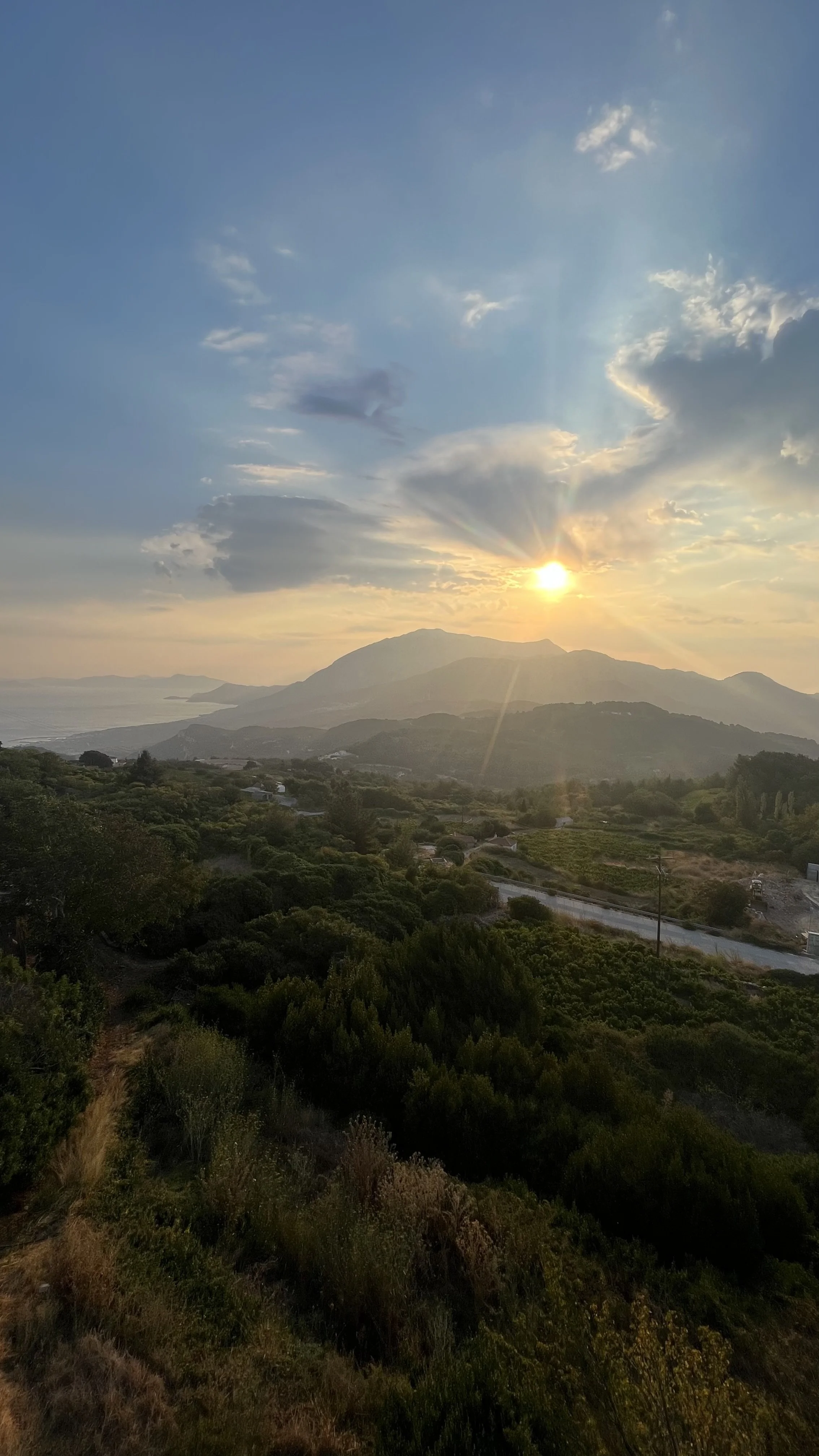 A scenic landscape at sunset with mountains in the distance, lush green trees and bushes covering rolling hills, and a sky with scattered clouds illuminated by the setting sun.