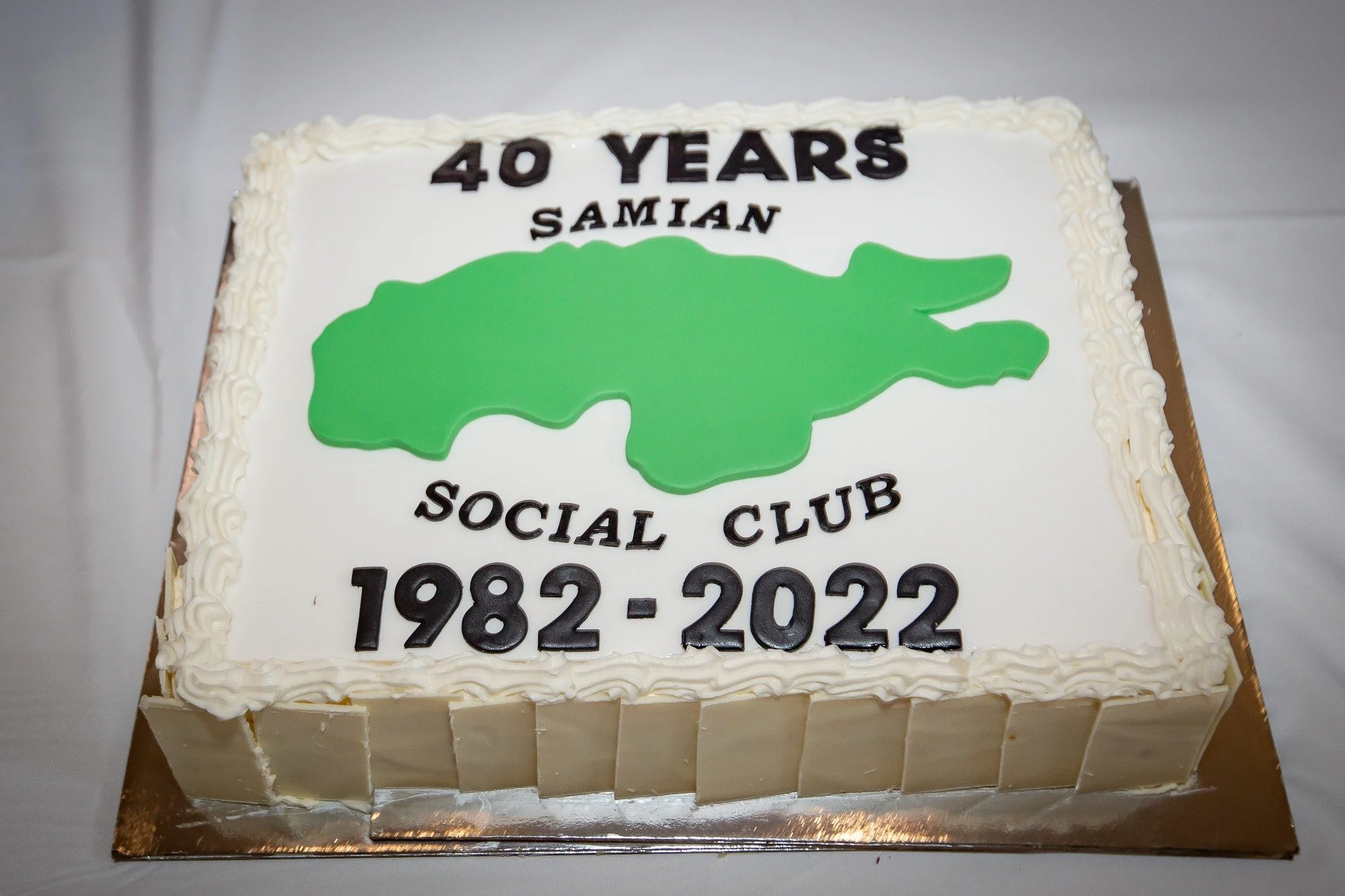 A rectangular celebration cake with white icing and piped border, featuring green graphic of a crocodile and text commemorating 40 years of Saman Social Club from 1982 to 2022.
