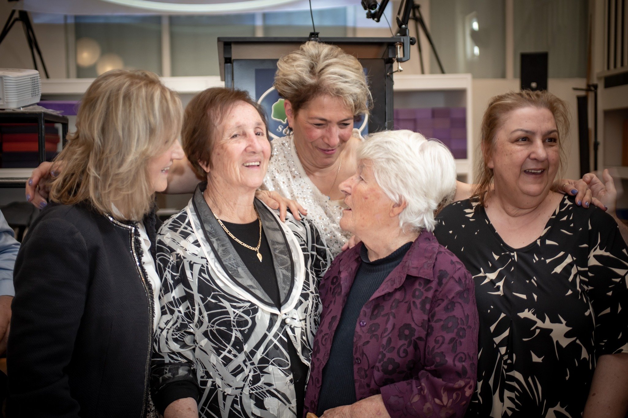 A group of five women, most likely senior citizens, are in a close, happy embrace and appearing joyful, in an indoor setting with background equipment and lighting.