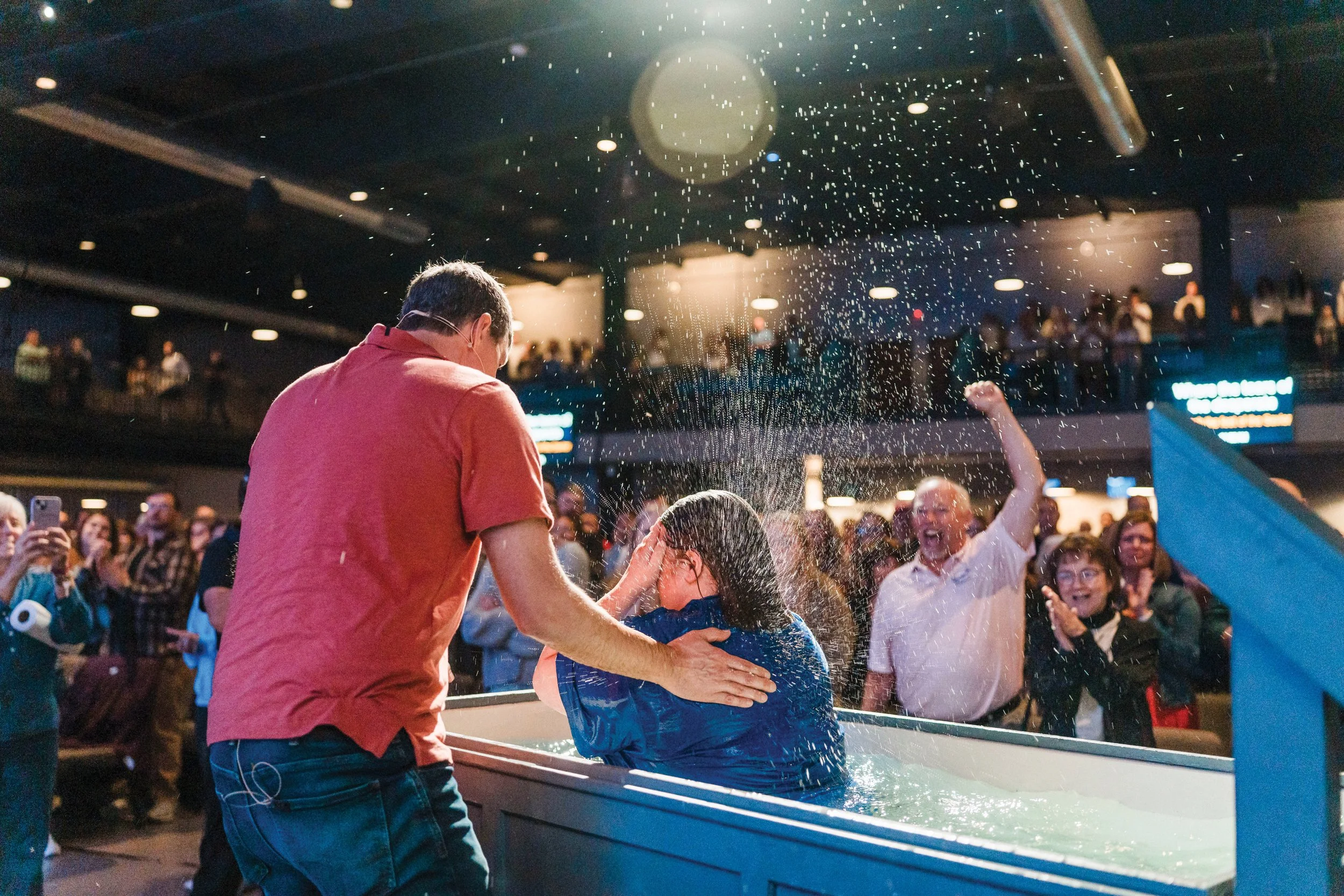 Pastor Glenn Greiner baptizes a parishioner who is coming up from the baptismal font. A man is cheering in the background.