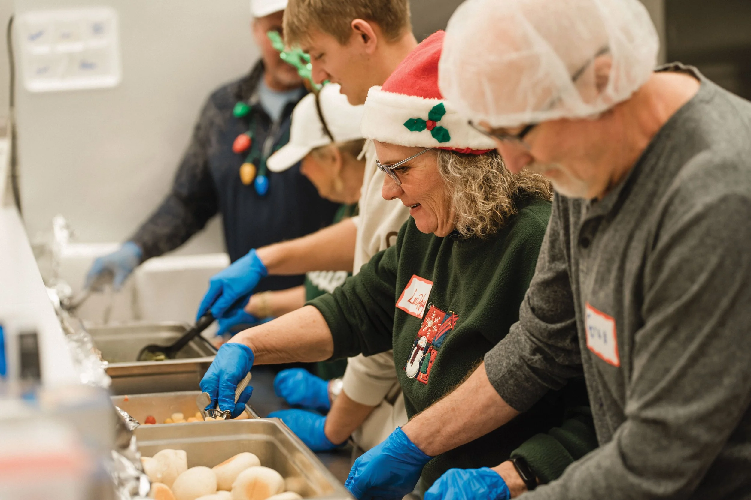Volunteers wearing hairnets and Christmas accessories serve food at a meal line.