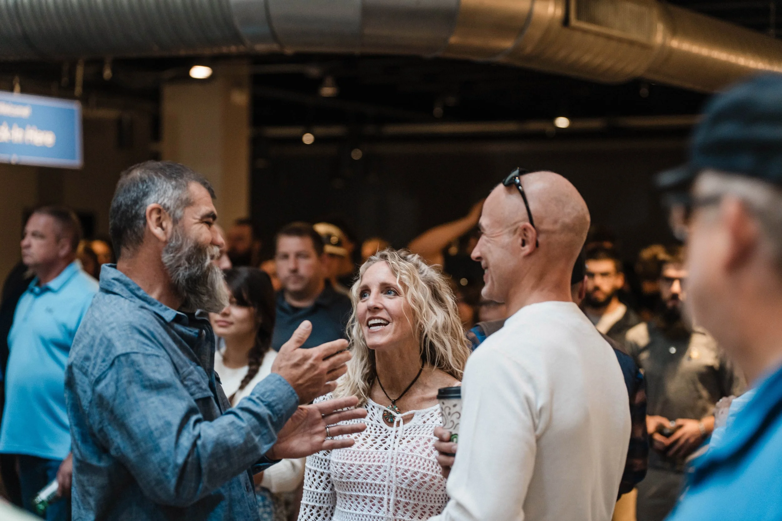 A group of two men and a woman smiling at each other converse in a crowded church lobby.