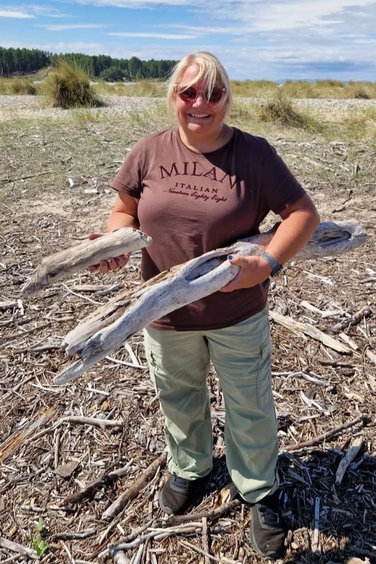 A woman with blonde hair and sunglasses, smiling and holding multiple pieces of driftwood on a sandy, debris-covered beach with grassy dunes and a line of trees in the background.