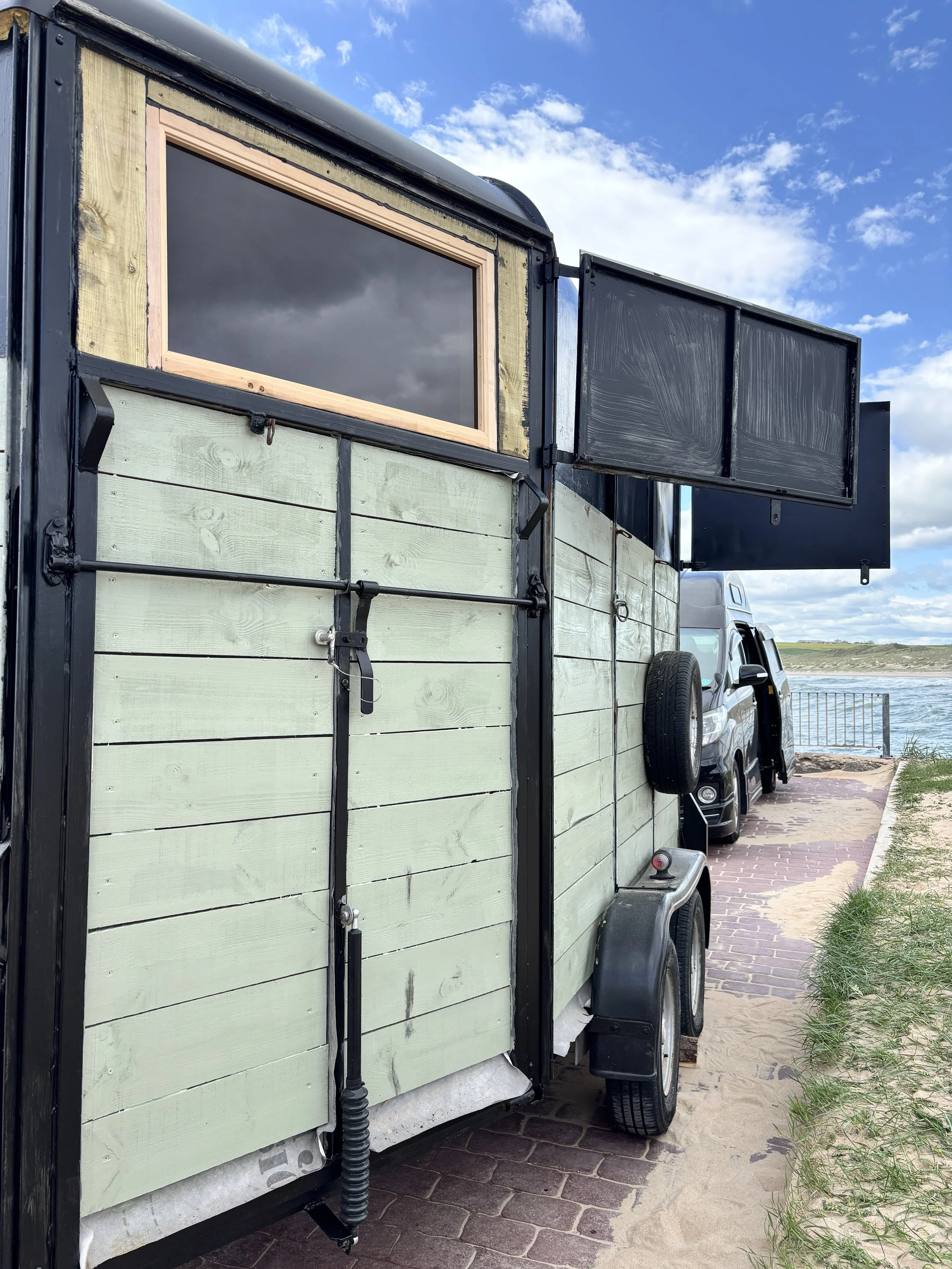 A trailer with a light green wooden exterior and black trim, parked beside a waterfront with a paved walkway and grassy area, under a partly cloudy sky.