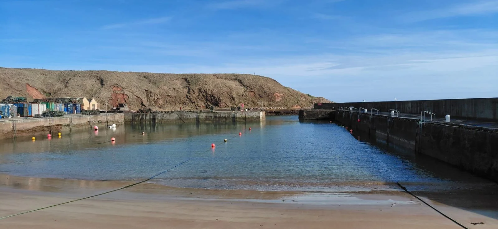 Port Erroll harbor with calm water, surrounded by a concrete pier and buoy markers, with a hillside in the background under a blue sky.