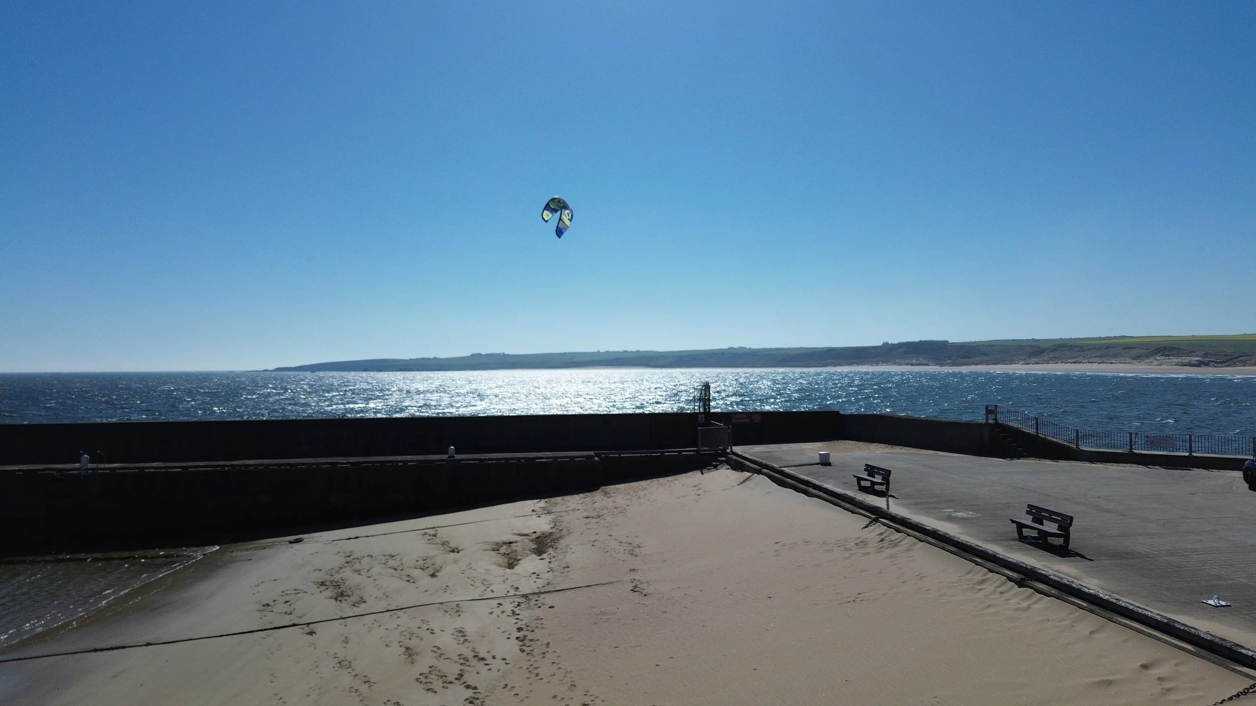 Beachside view with a concrete promenade, benches, and railings overlooking the ocean under a clear blue sky, with a kite surfer in the distance.