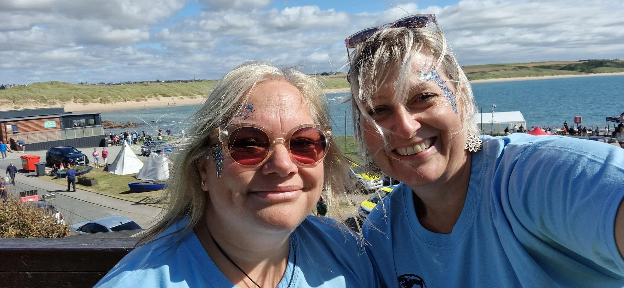 Two women with blonde hair smiling, wearing light blue shirts, at a seaside event with tents, people, and vehicles in the background on a partly cloudy day.