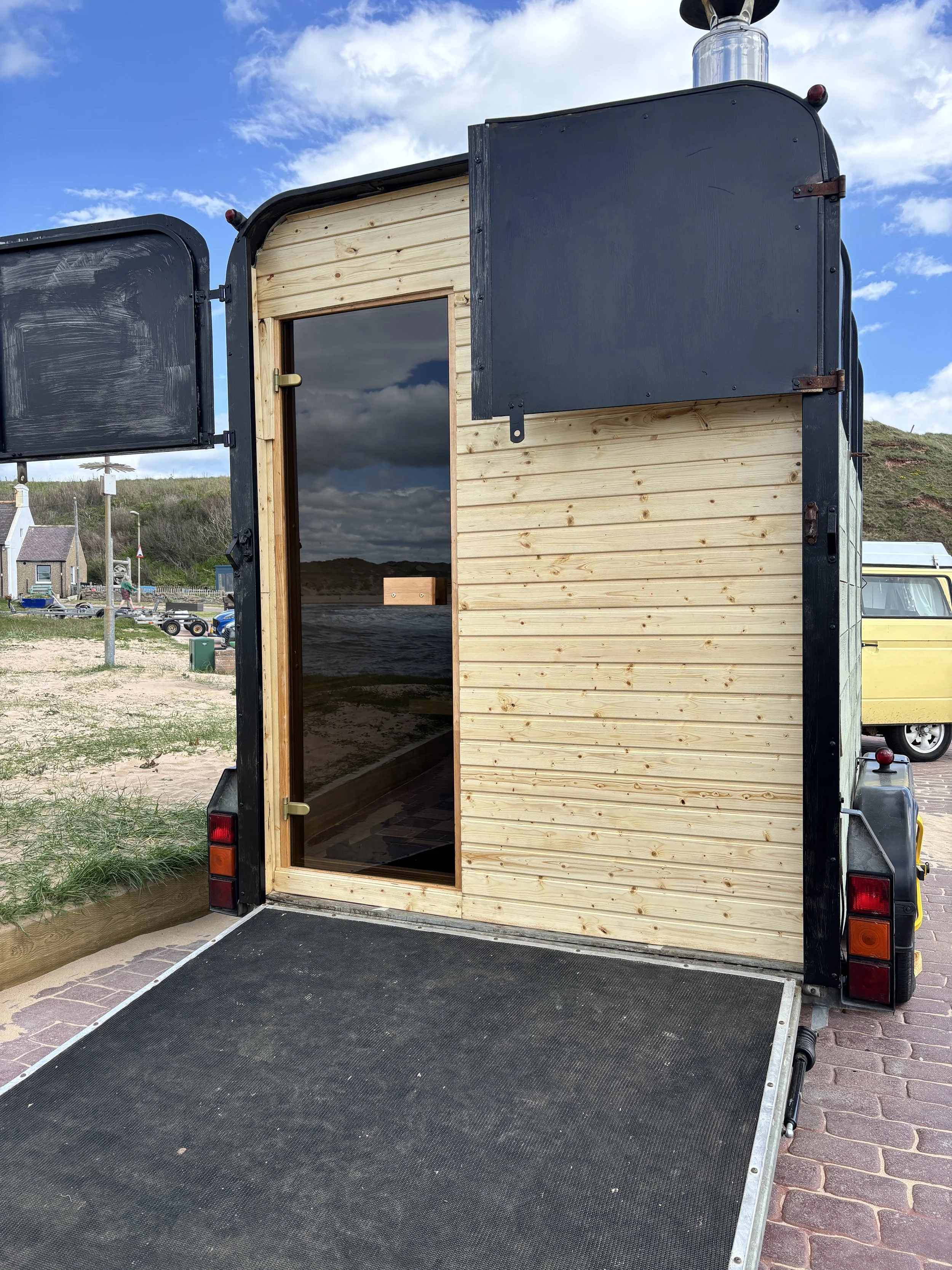 The door to the wood-fired sauna.  The glass door shows a wooden interior, on a paved surface outdoors, with a blue sky and clouds overhead.