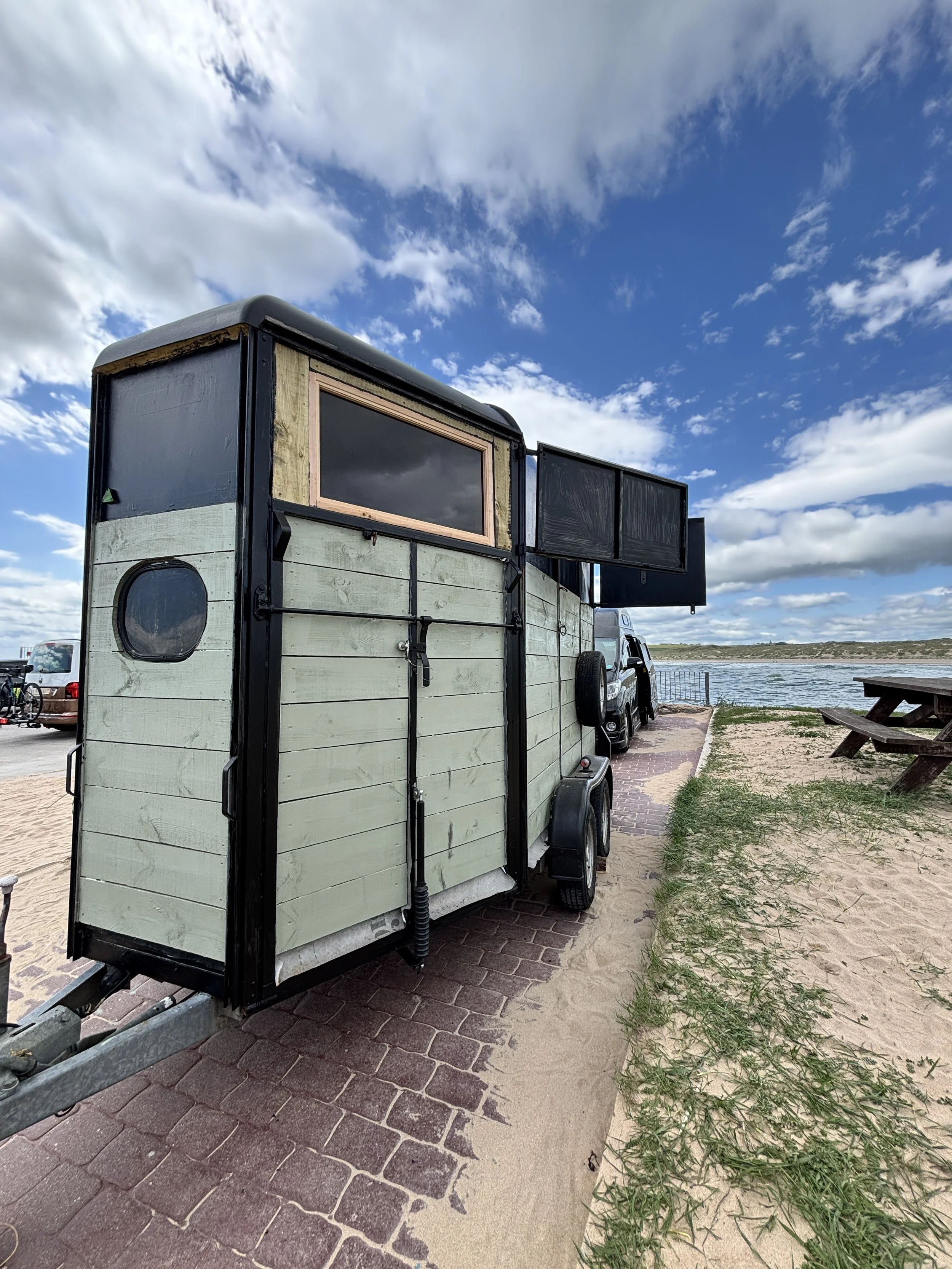 A converted horsebox sauna parked near a body of water, with a picnic table nearby, under a partly cloudy sky.