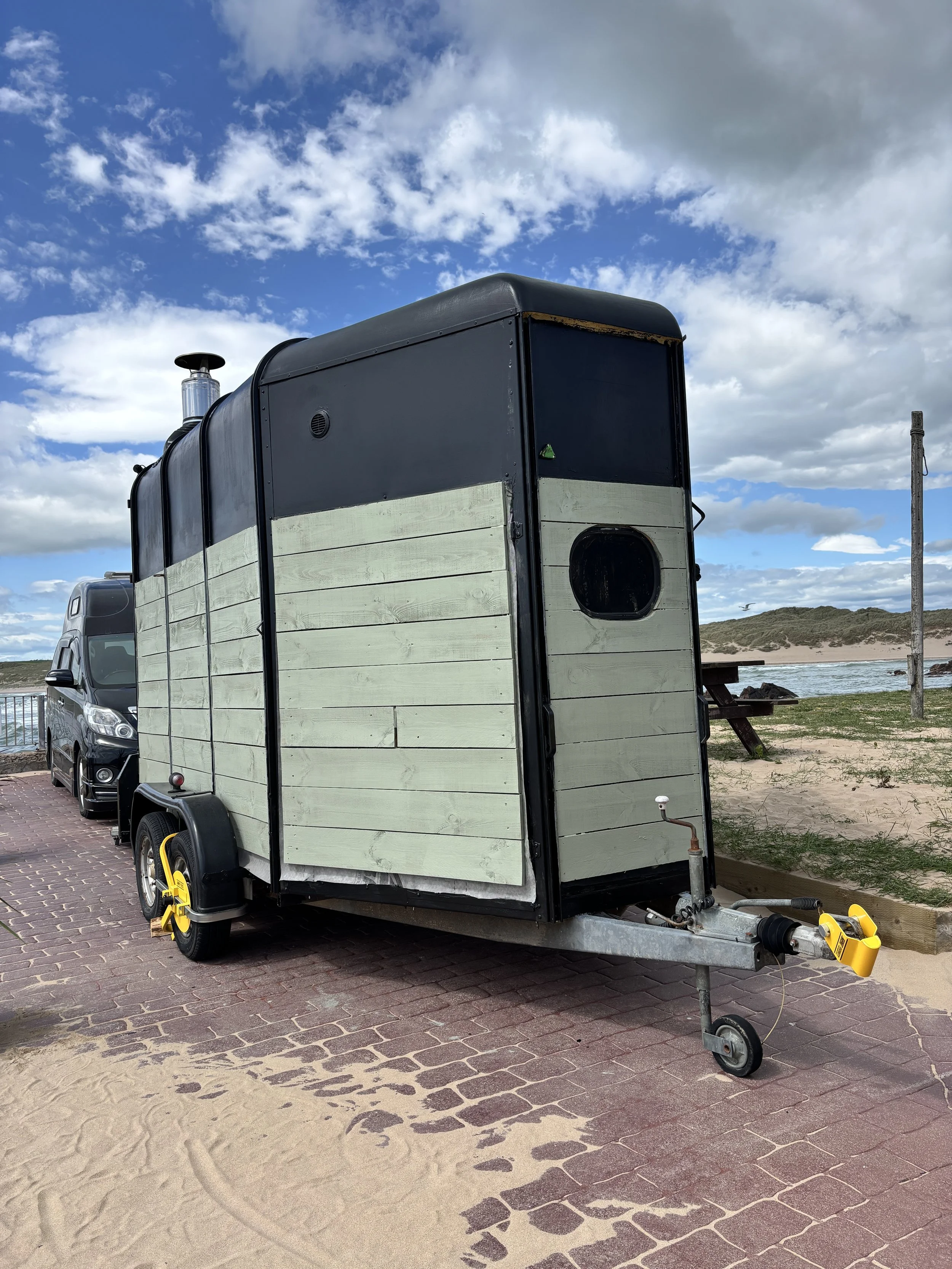 A converted horsebox.  A wood-fired sauna with a sage green wooden exterior and black trim parked on a paved path near the beach. In the background, the sandy beach, ocean, and a cloudy blue sky are visible.