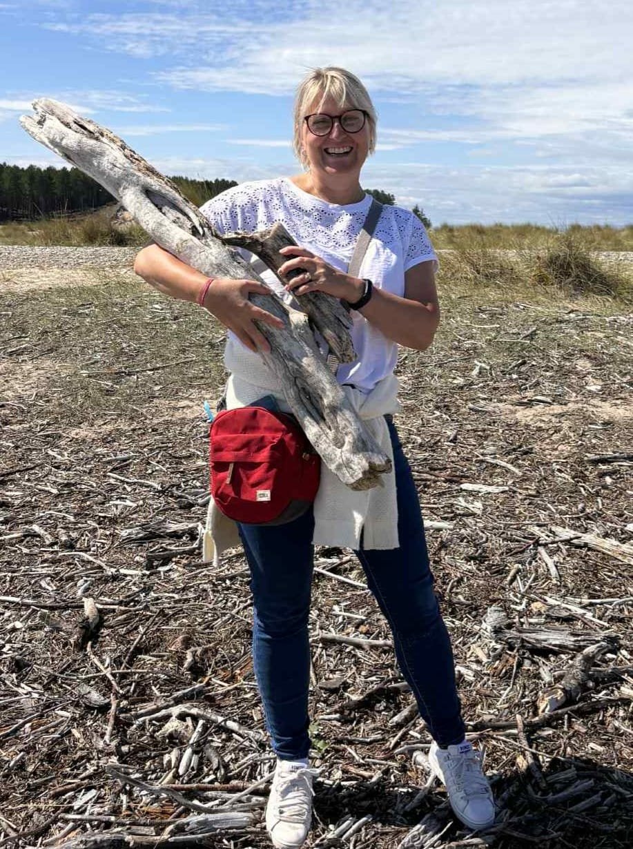 Woman in casual clothes and glasses holding large weathered driftwood on a sandy, log-strewn beach with grassy dunes and blue sky in the background.