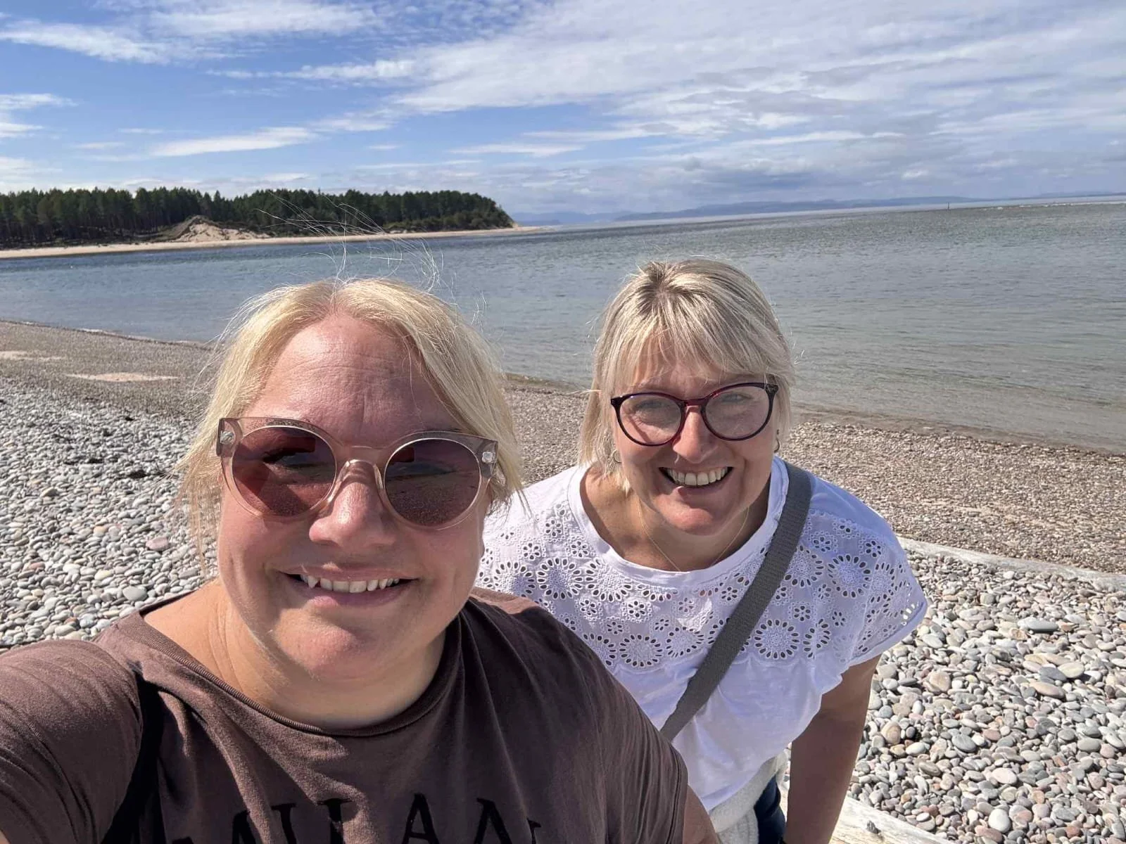 Two women smiling at the beach, with pebbles, water, and green trees in the background, under a partly cloudy sky.