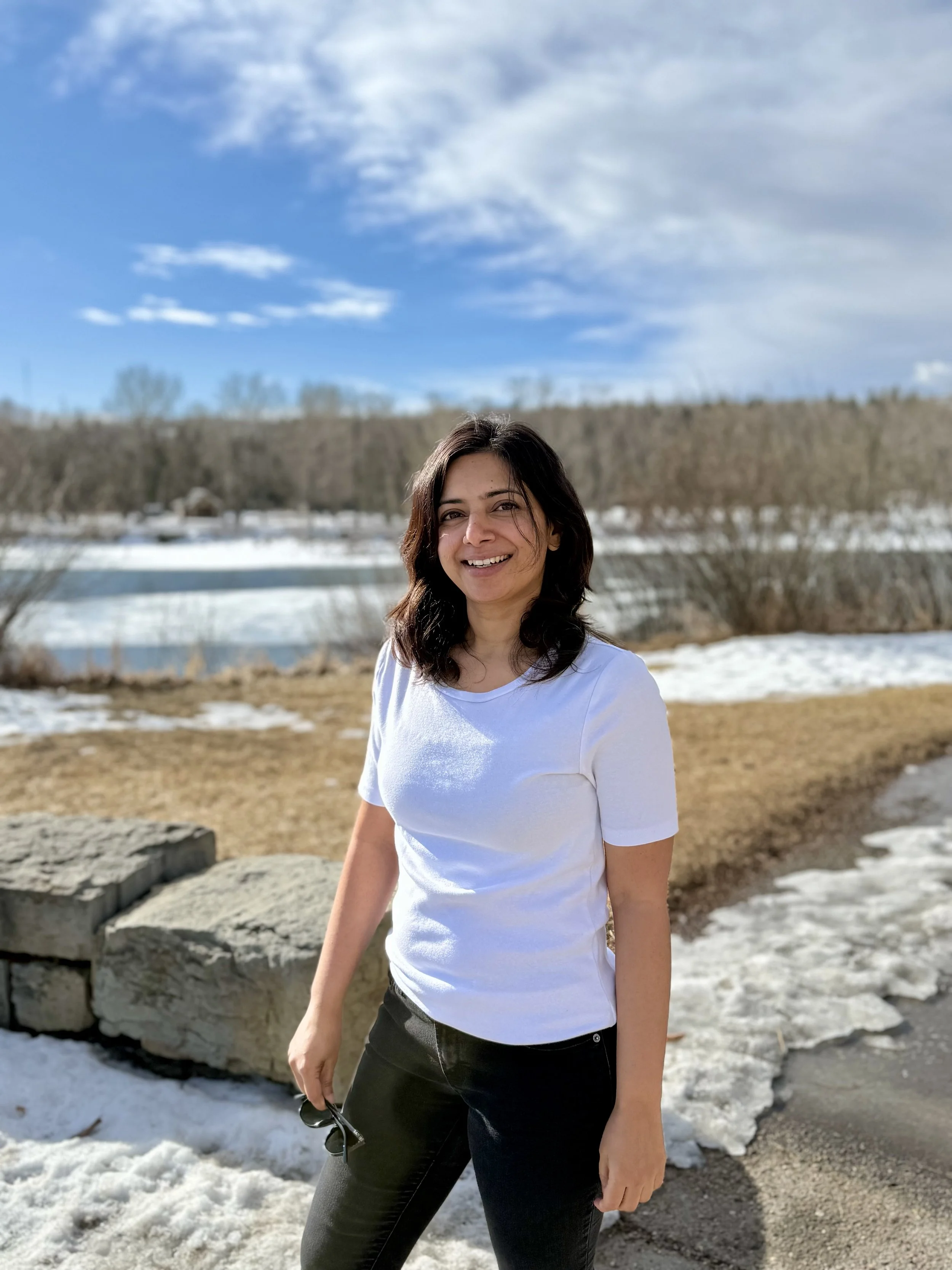 A smiling woman with dark hair wearing a white t-shirt and black pants standing outdoors on a partially snowy landscape with a river, leafless trees, and a blue sky with white clouds in the background.