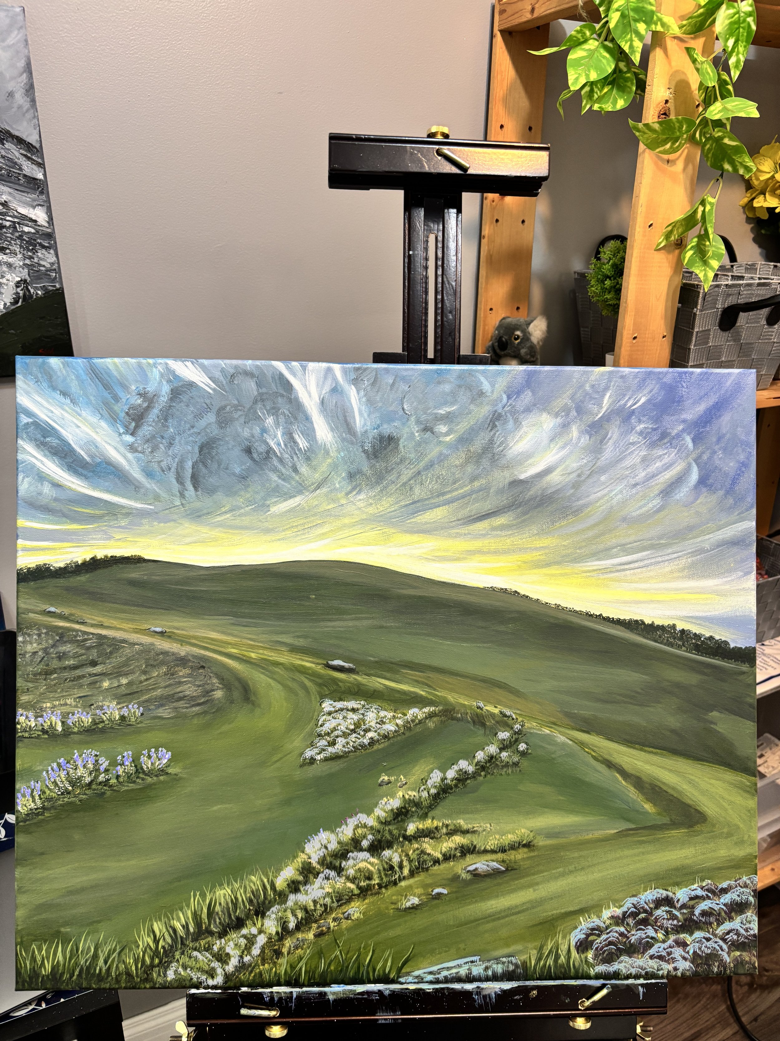 A landscape painting depicting rolling green hills with patches of purple and white flowers under a dramatic sky with swirling clouds at sunset.