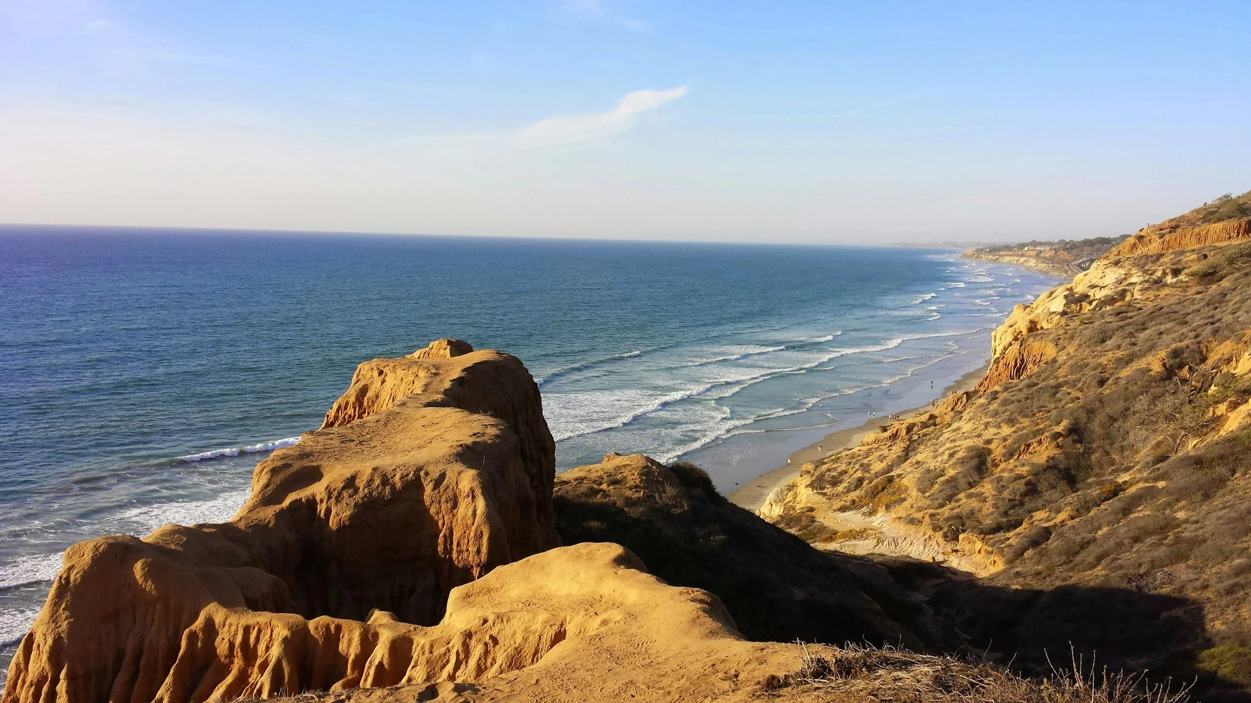 A scenic view of a sunset over the ocean with rocky cliffs in the foreground and a sandy beach stretching along the coastline.