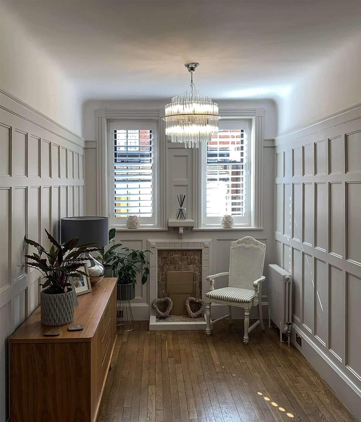 A cozy, bright living room with white paneled walls, two large windows with shutters, a chandelier, a white armchair, a wooden sideboard with decorative plants and a picture frame, a small fireplace with heart-shaped decorations, and hardwood flooring.