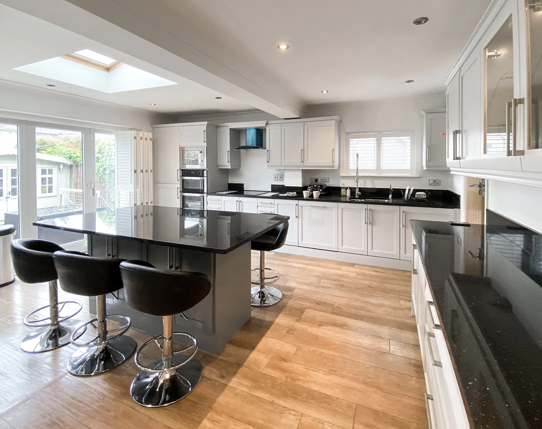 Modern kitchen with white cabinets and black countertops, a central island with black chairs, and large windows with shutters, hardwood flooring, and skylight ceiling.