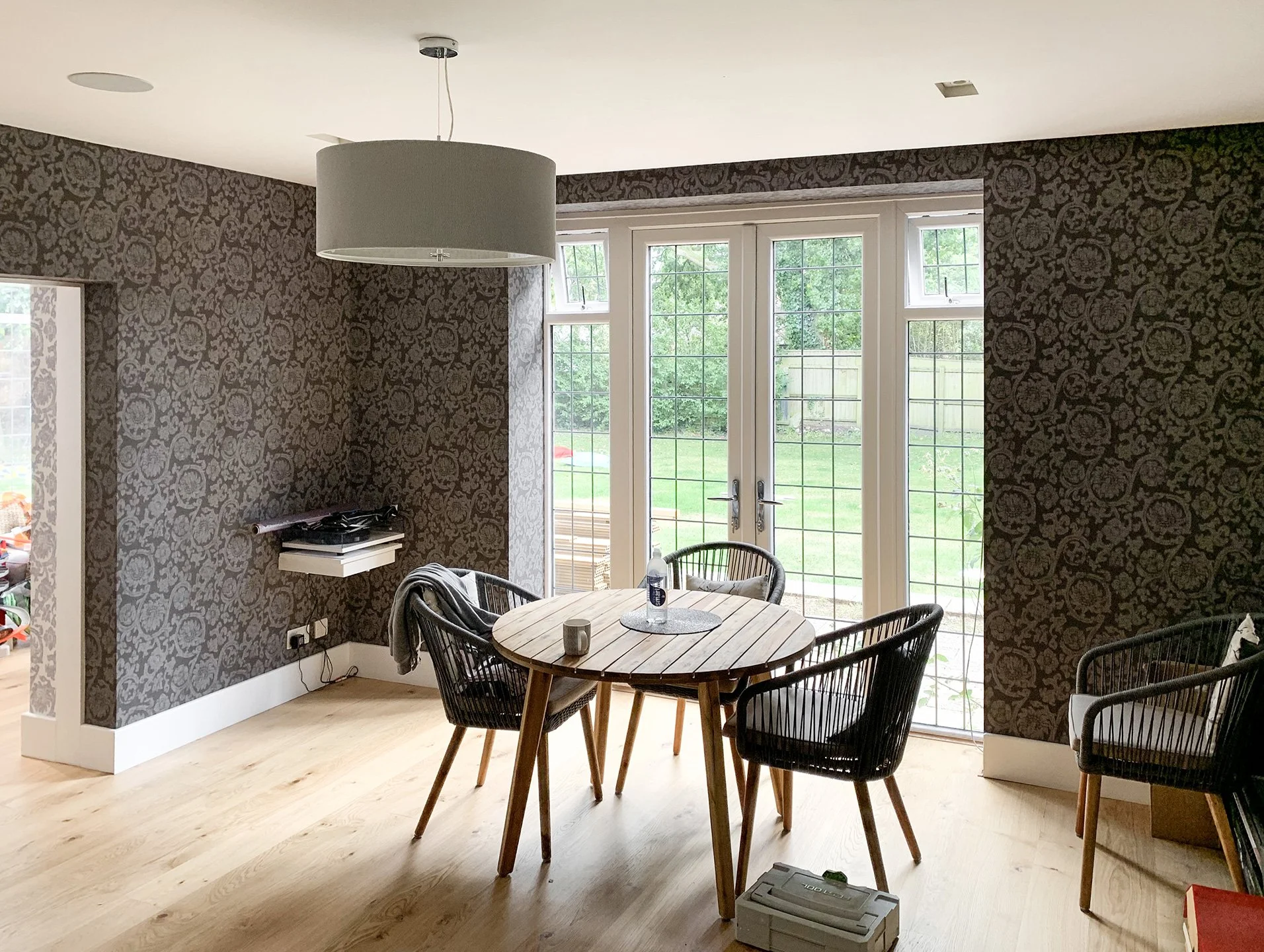 A dining area with a round wooden table surrounded by black wire chairs, a large window with a sliding glass door looking out to a backyard, and brown patterned wallpaper on the wall.