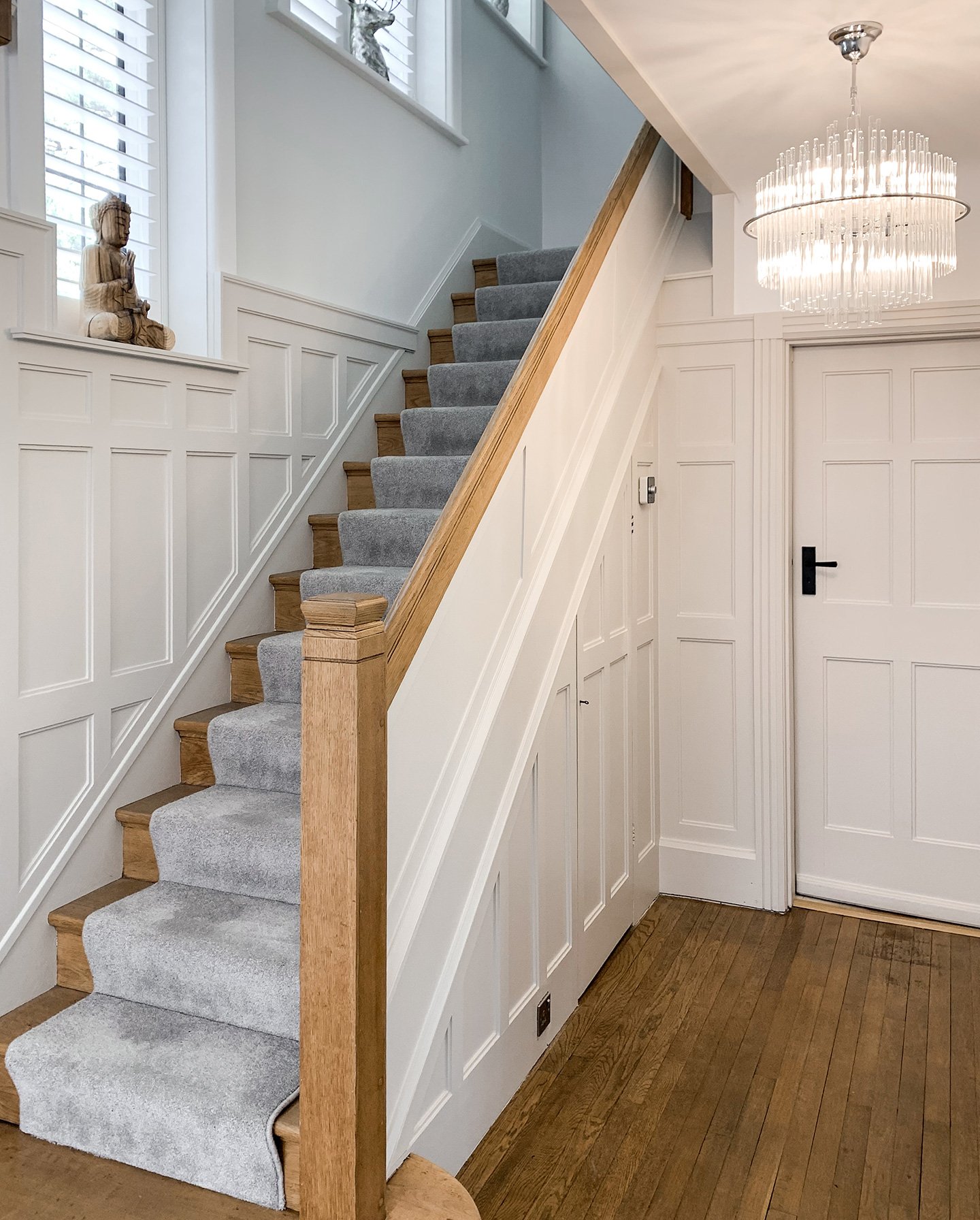 Interior view of a staircase with wooden steps, gray carpet runner, white paneled walls, and decorative decor on the wall and window ledges. A chandelier hangs from the ceiling near a white door.