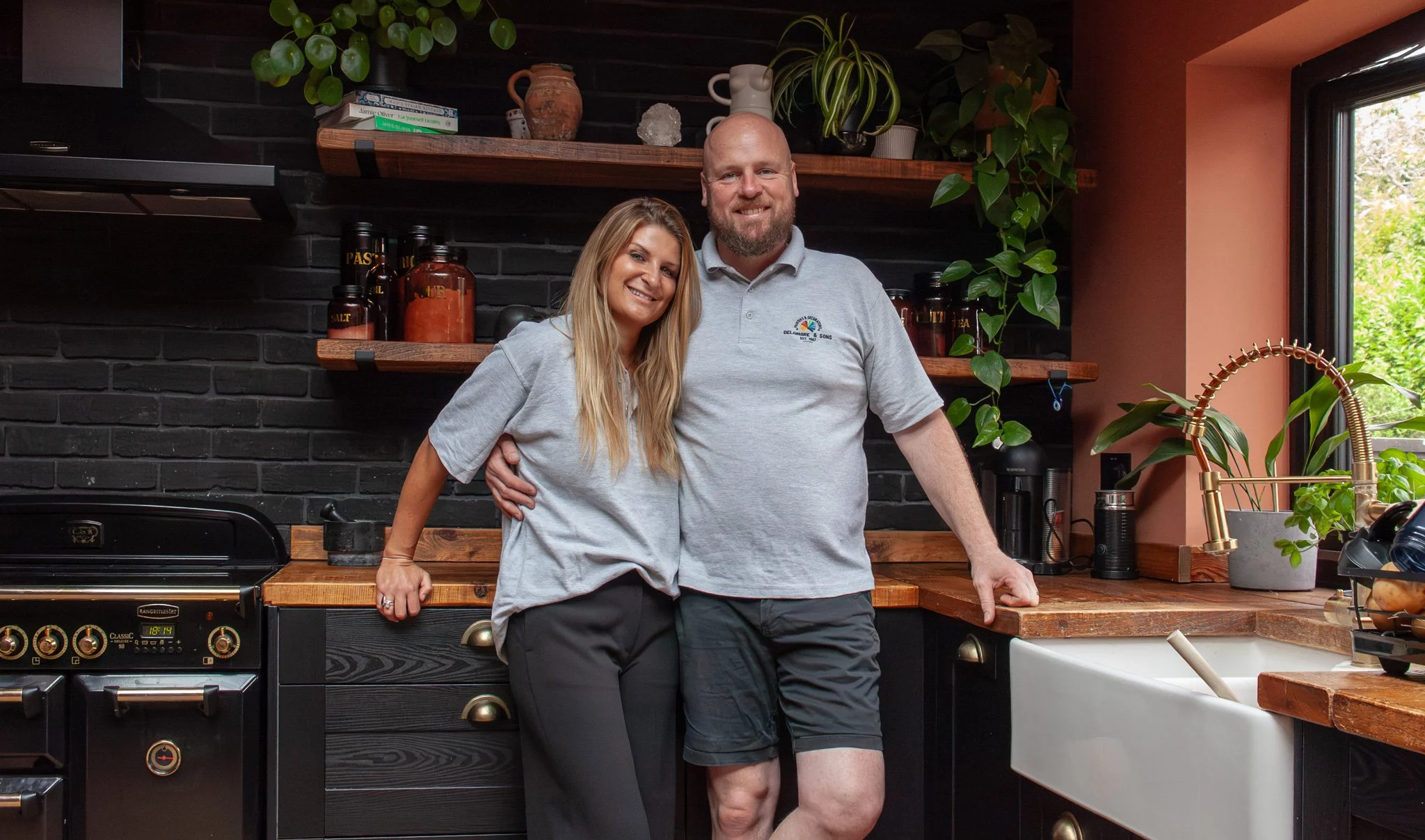 A smiling couple stands together in a modern kitchen next to a black stove. The woman has long blonde hair and is wearing a gray shirt, and the man has a shaved head and beard, wearing a light gray polo shirt. There are green plants and kitchen items on the shelves behind them, and a window to the right lets in natural light.