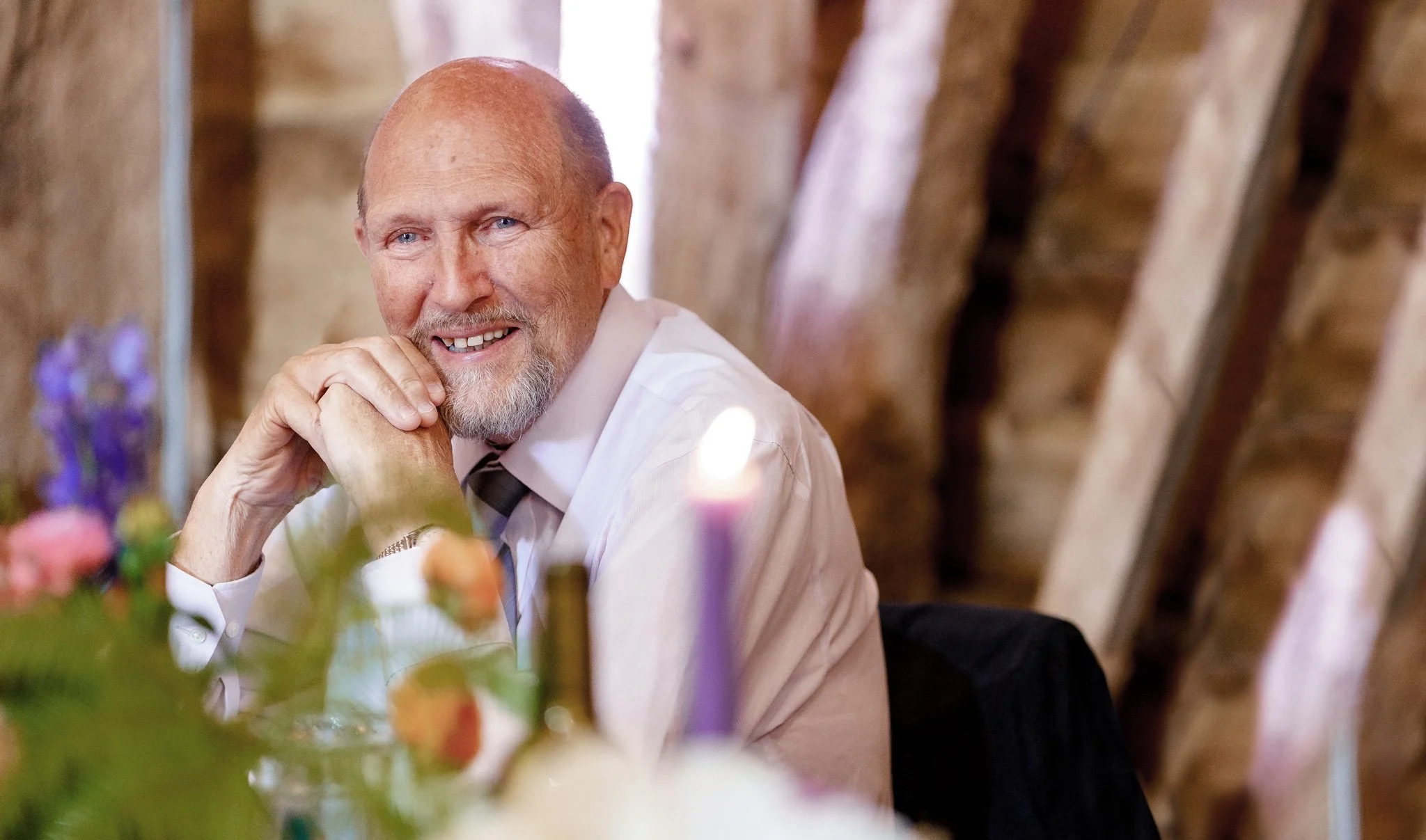 A smiling middle-aged man with a beard and blue eyes, wearing a light pink shirt and gray tie, sitting at a table with colorful flowers and soft lighting in the background.