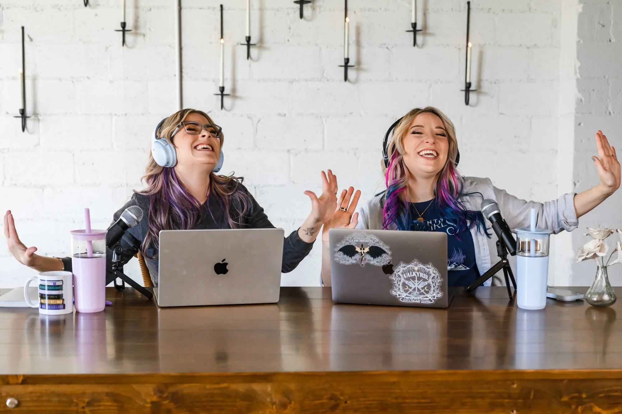 Two women with colorful hair and headphones laughing and recording a podcast at a wooden table with laptops and microphones.