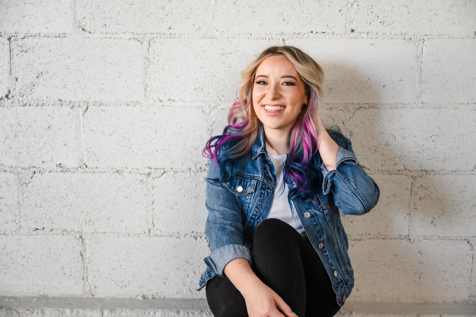 A young woman with multi-colored hair smiling and sitting against a white brick wall, wearing a denim jacket and black pants.