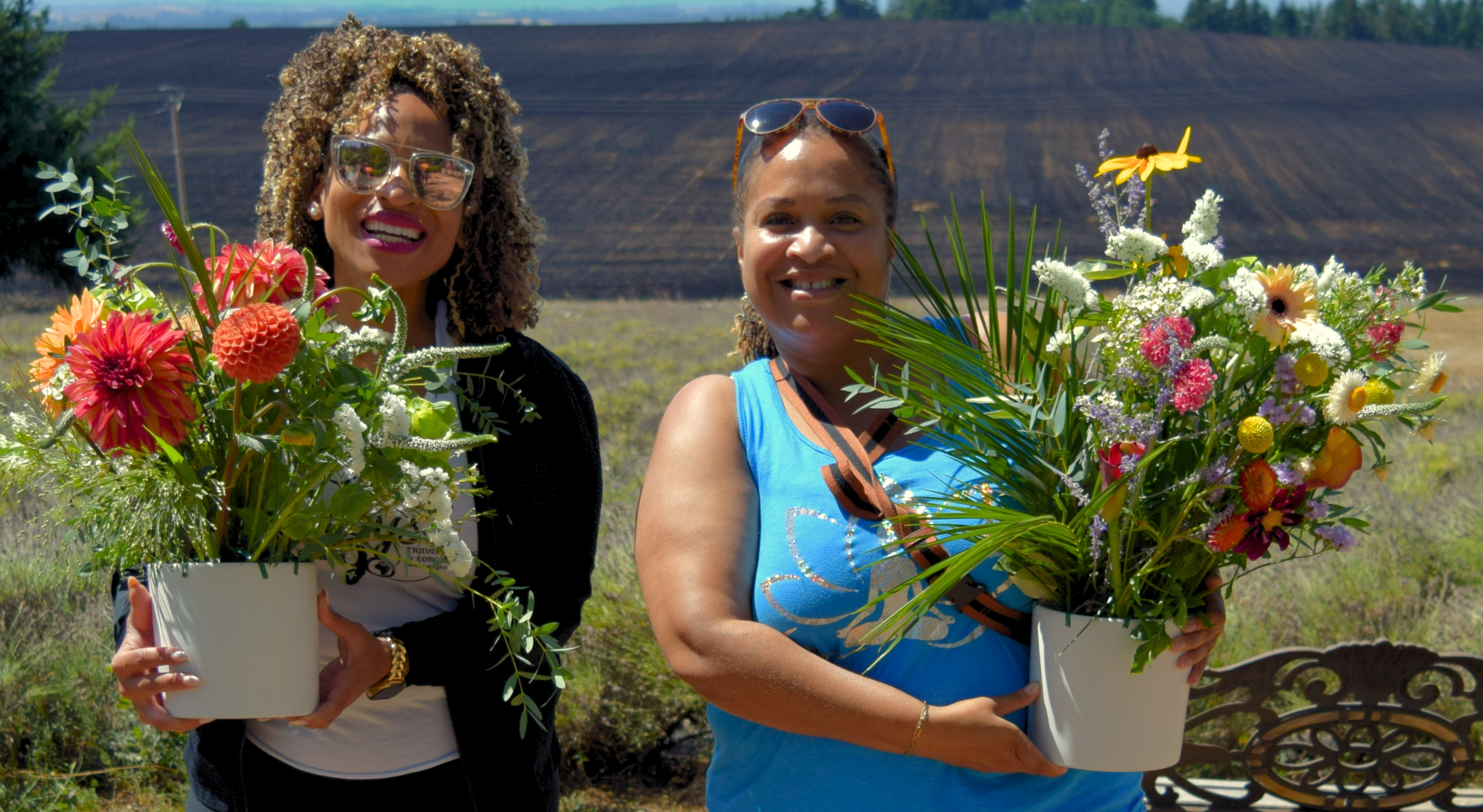 Two women leaders outdoors holding large white pots filled with colorful flowers, with open fields and a hill in the background.