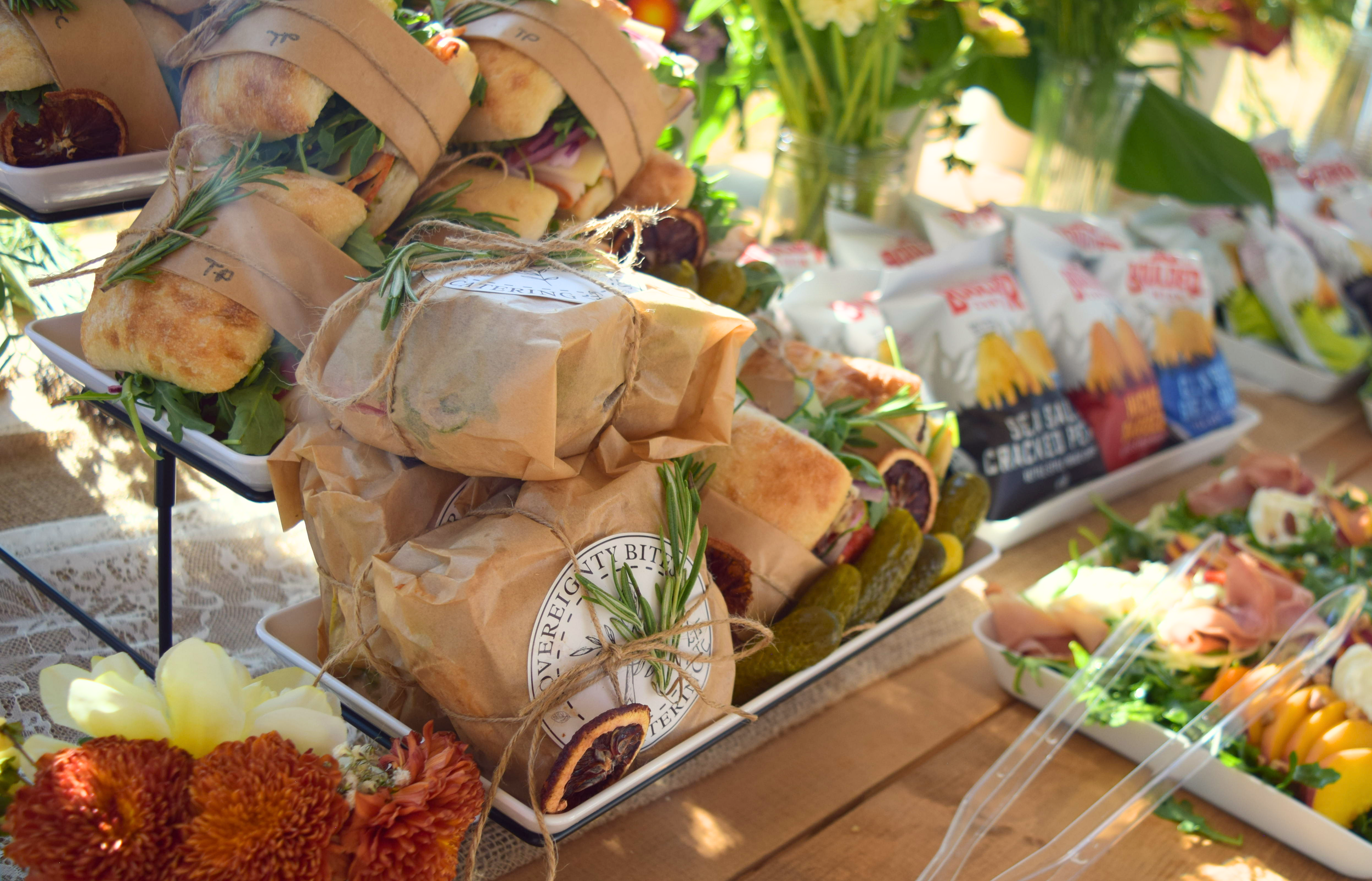 A display of wrapped sandwiches, snacks, and salads on a wooden table, decorated with flowers and greenery, with sunlight filtering through the background.