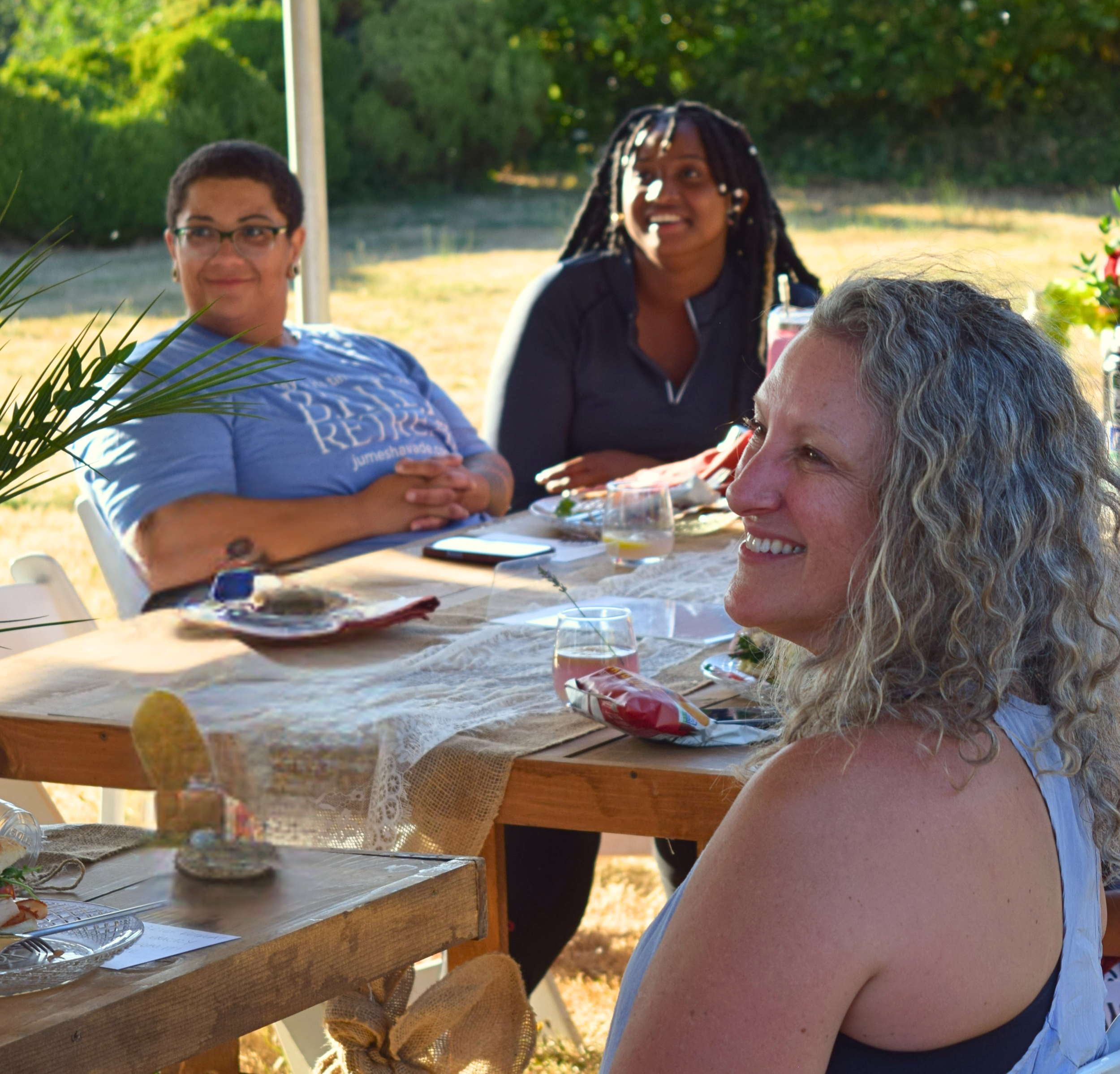 Three women leaders and collaborators sitting at a dining table outdoors, smiling and enjoying a gathering on a sunny day with greenery in the background.