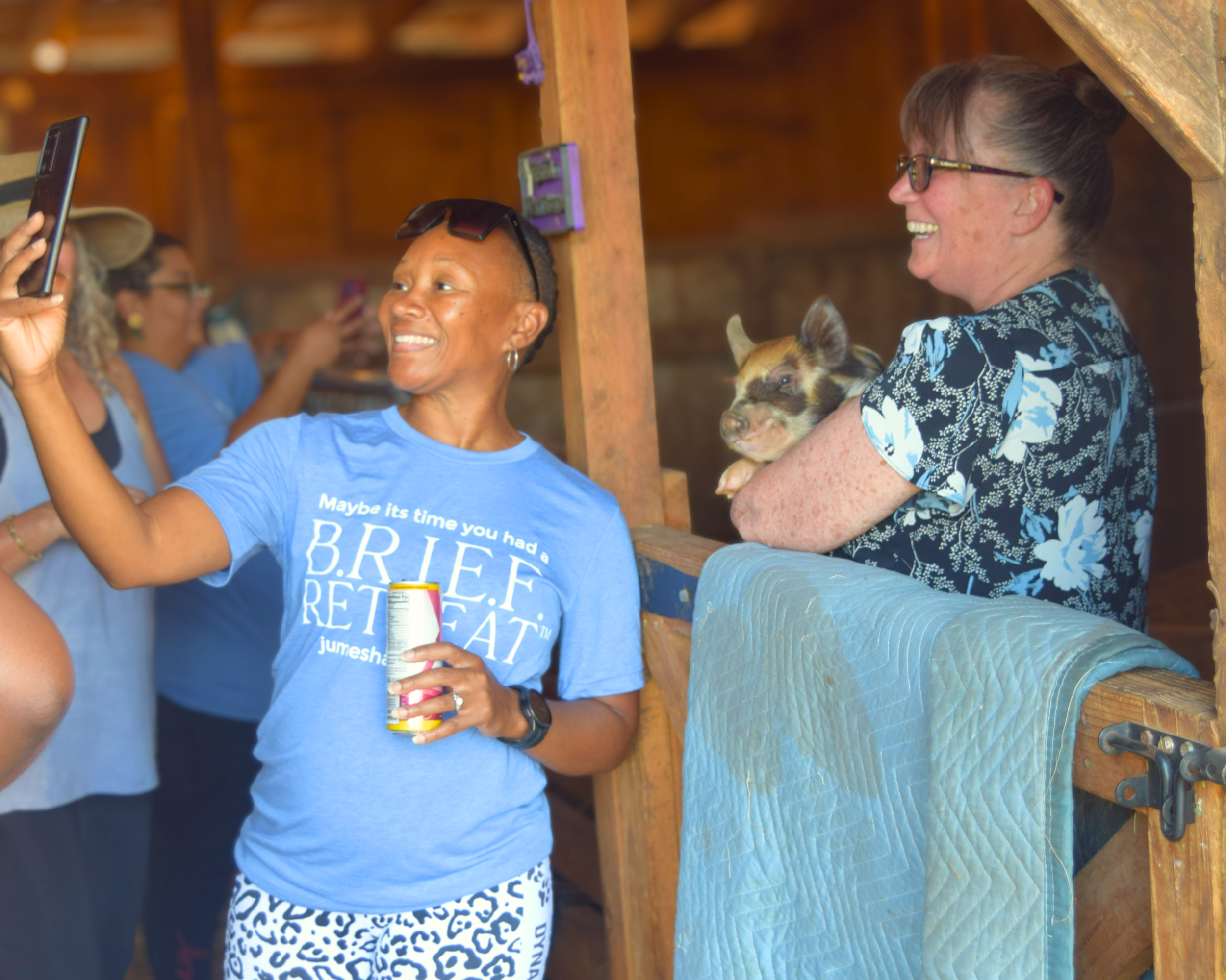 Two women in a barn, one taking a selfie and the other holding a piglet, both smiling.