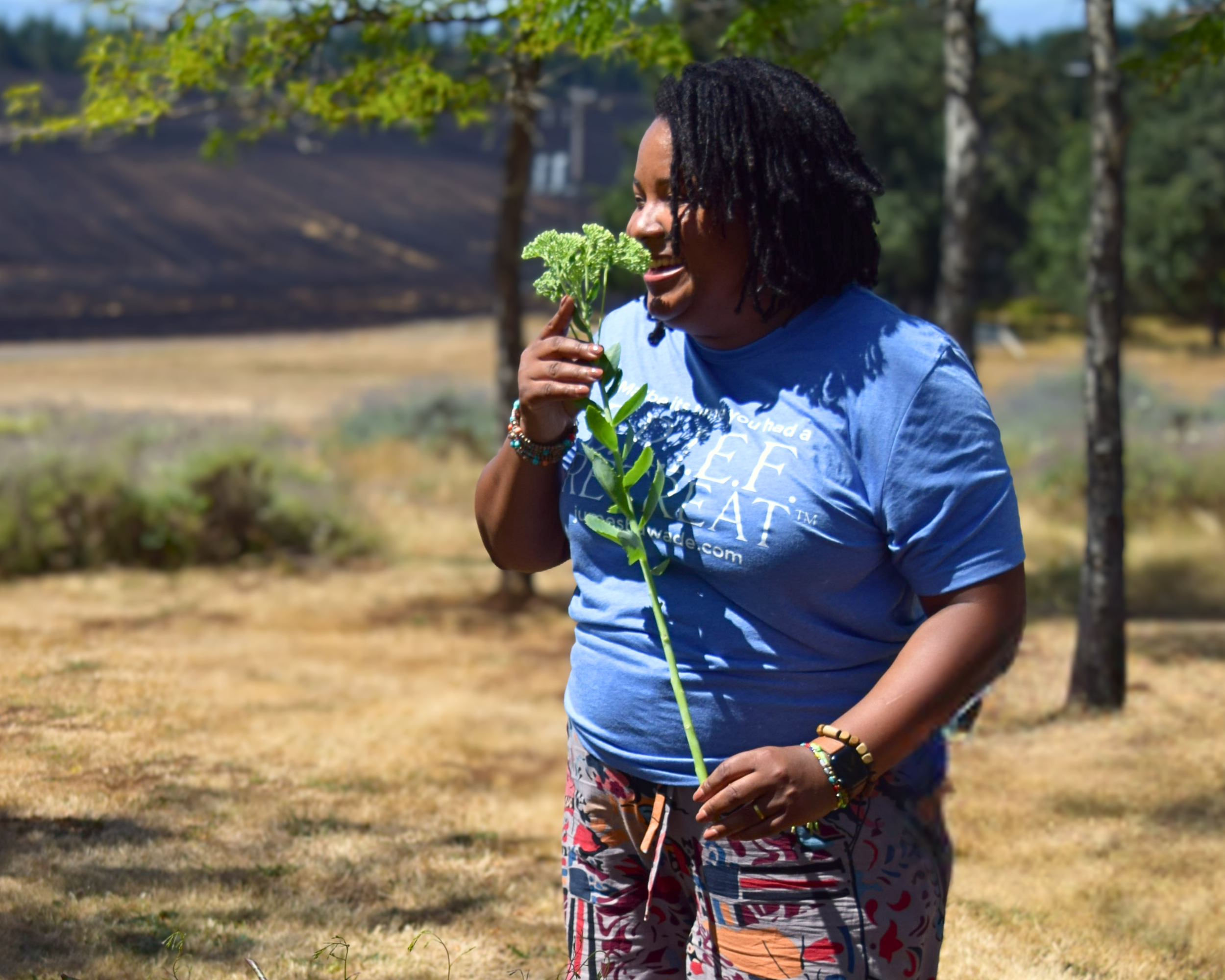 A retreat leader smiling outdoors, holding a green leafy plant close to her face, wearing a blue T-shirt and colorful patterned pants, surrounded by trees and natural landscape - preparing to speak with attendees.