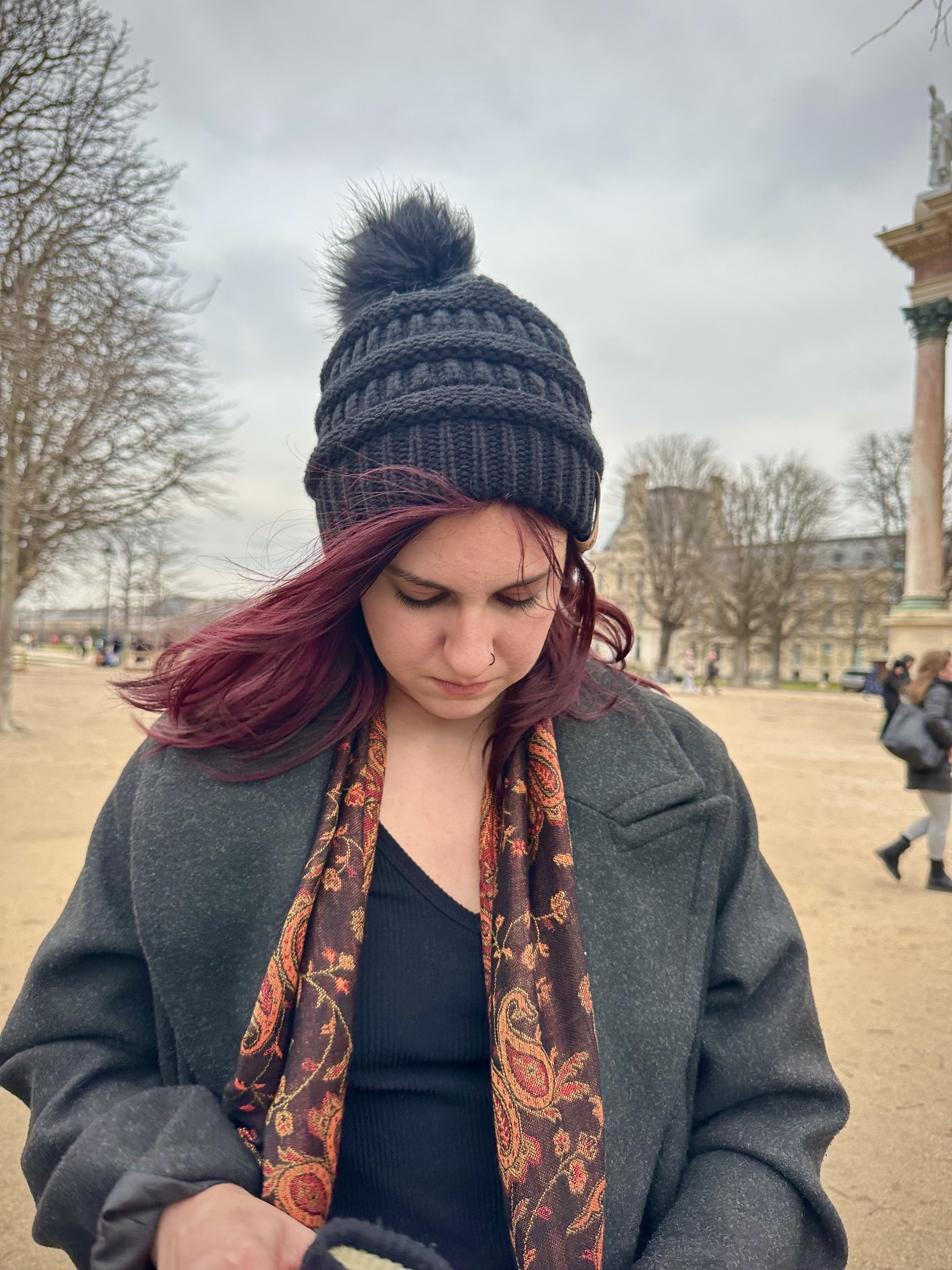 A young woman with maroon hair wearing a knit black beanie with a pom-pom, a dark coat, and a patterned scarf, standing outdoors in a park on a cloudy day.