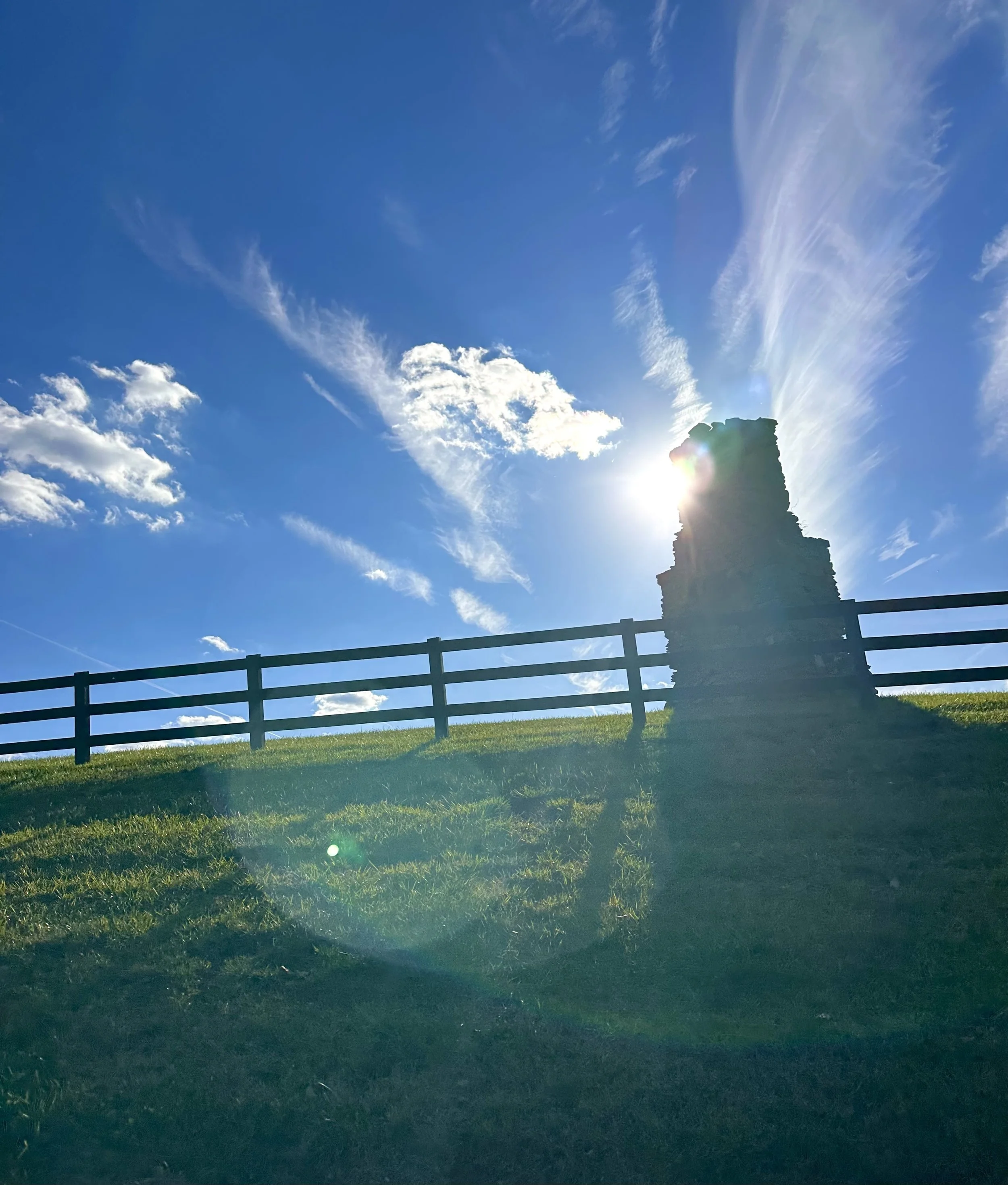 Sun shining behind an old chimney on a grassy hill with a black wooden fence and sky with clouds.