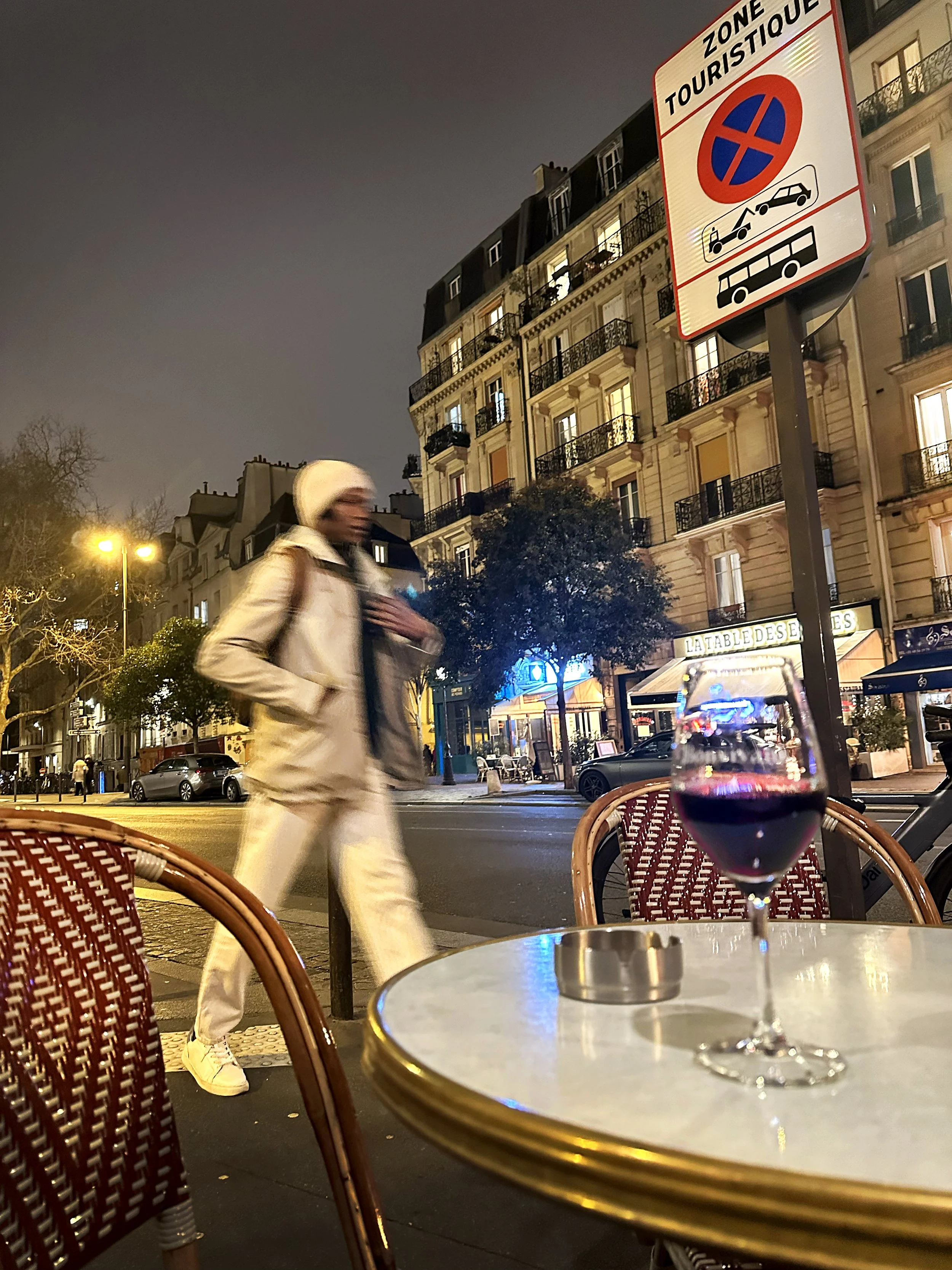 A street scene at night with a person walking past an outdoor café with a glass of red wine on the table. There are residential buildings in the background and a street sign indicating a tourist zone with restrictions on parking.
