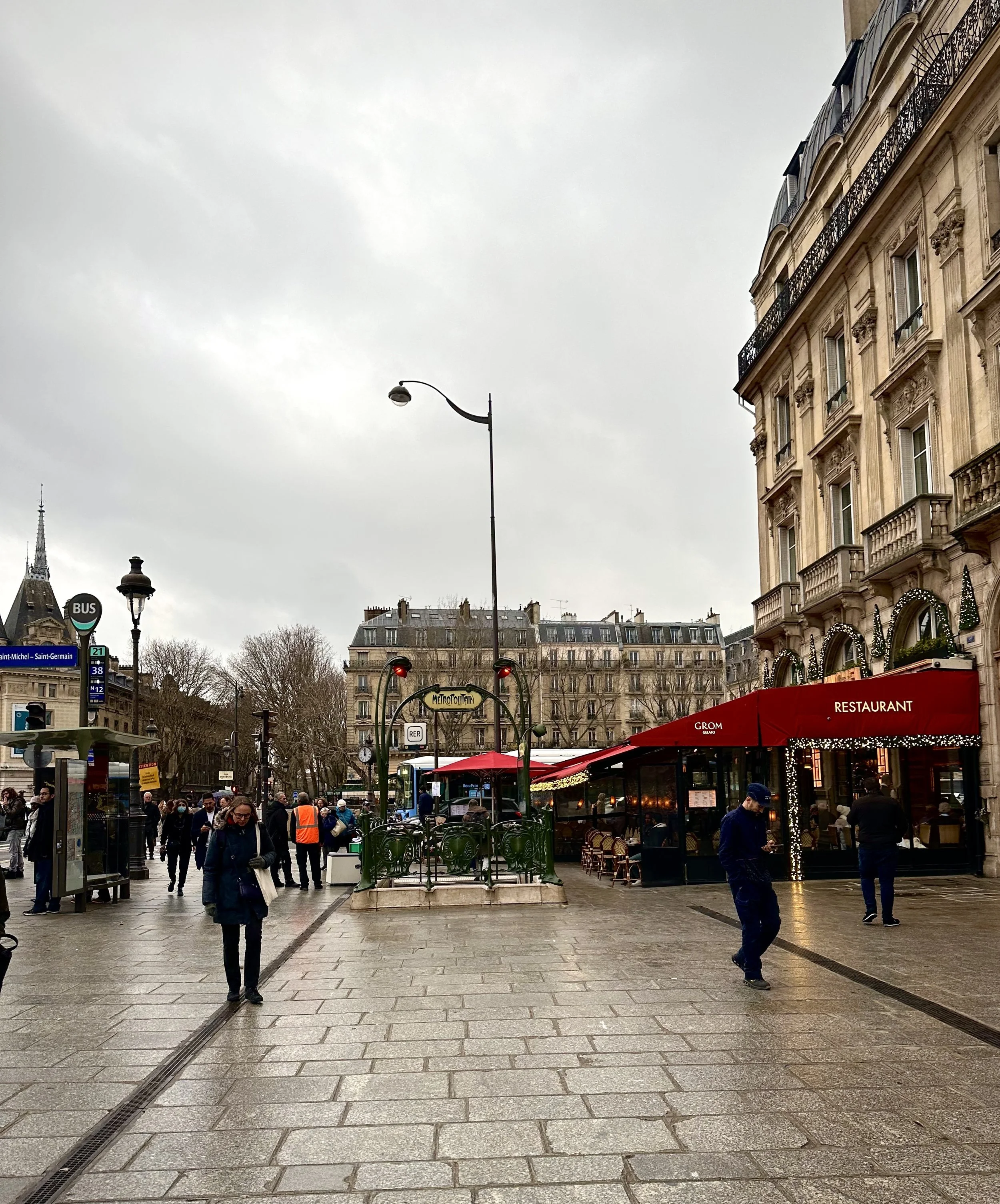 City street scene in Paris with people walking, a metro station entrance, a bus stop, and buildings with European architecture on a cloudy day.