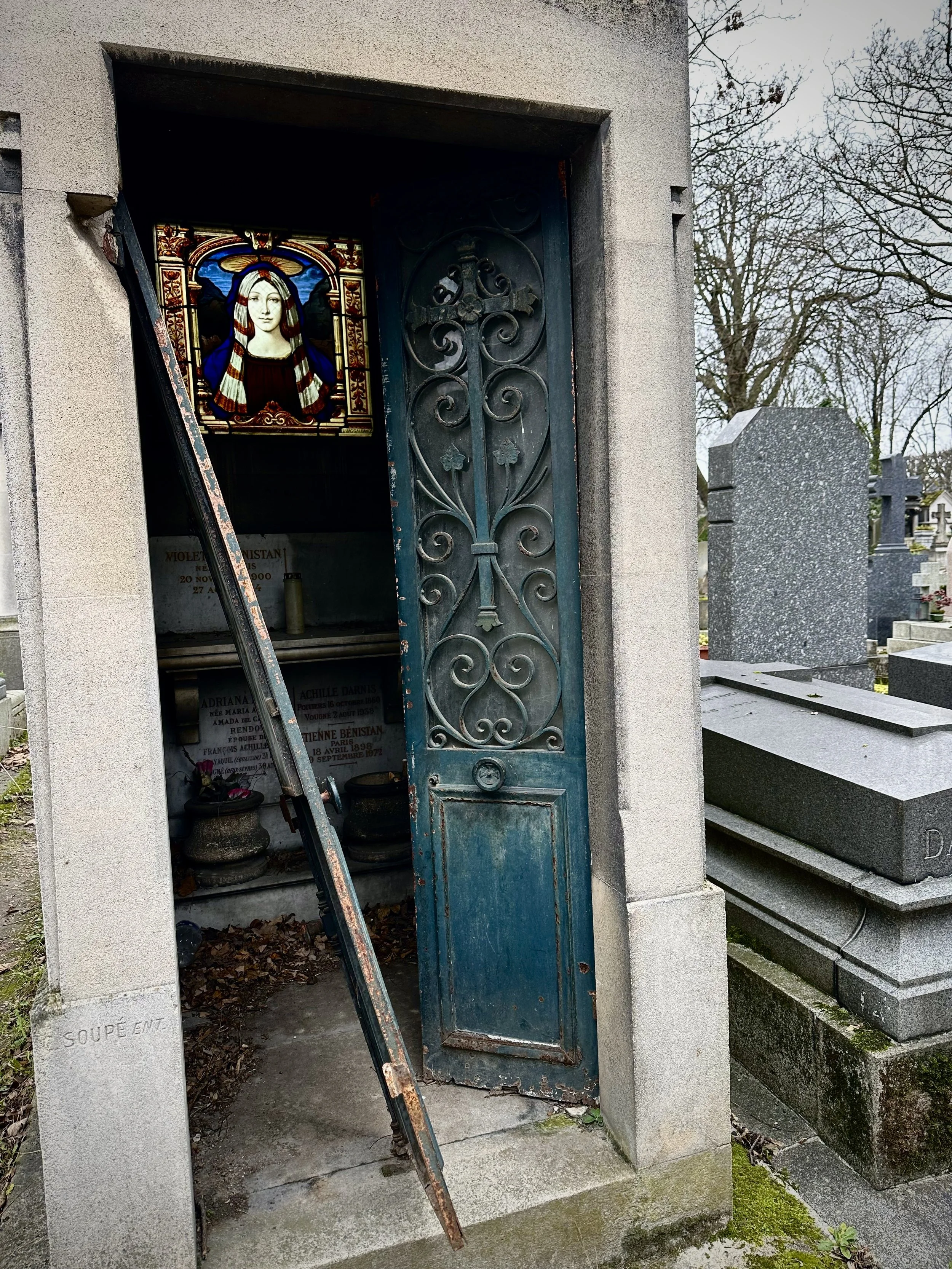A small tomb with an ornate iron door, inside a mausoleum or crypt, with a stained glass image of Mary and Jesus on the wall and gravestones in the background.