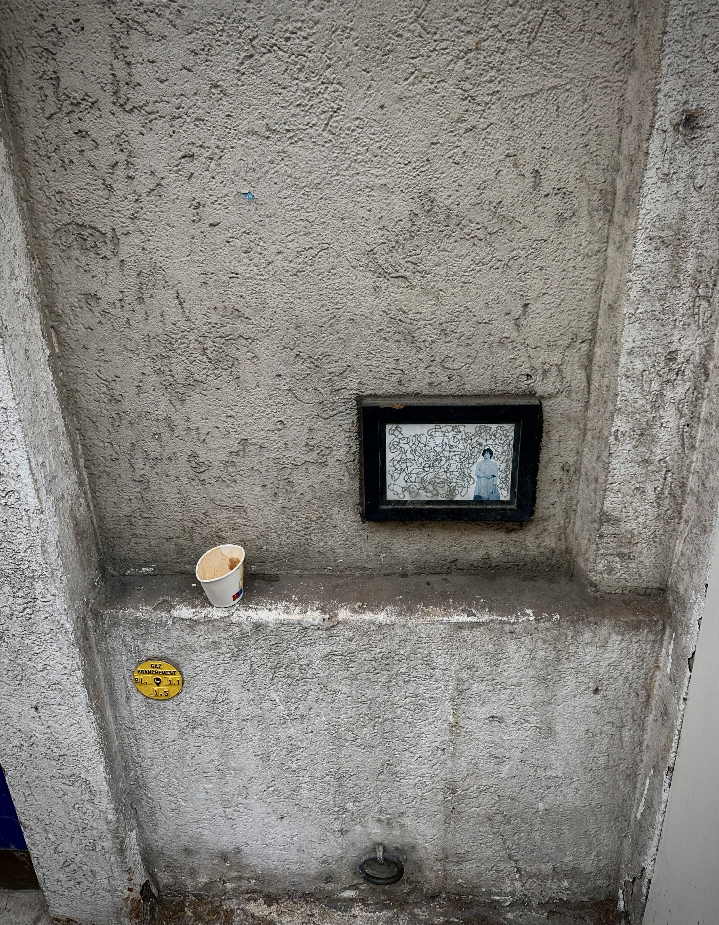 A textured, gray concrete wall with a small, clear framed picture of a woman standing amidst abstract patterns, a disposable coffee cup with a lid, a yellow gas meter label, and a metal pipe fitting below.