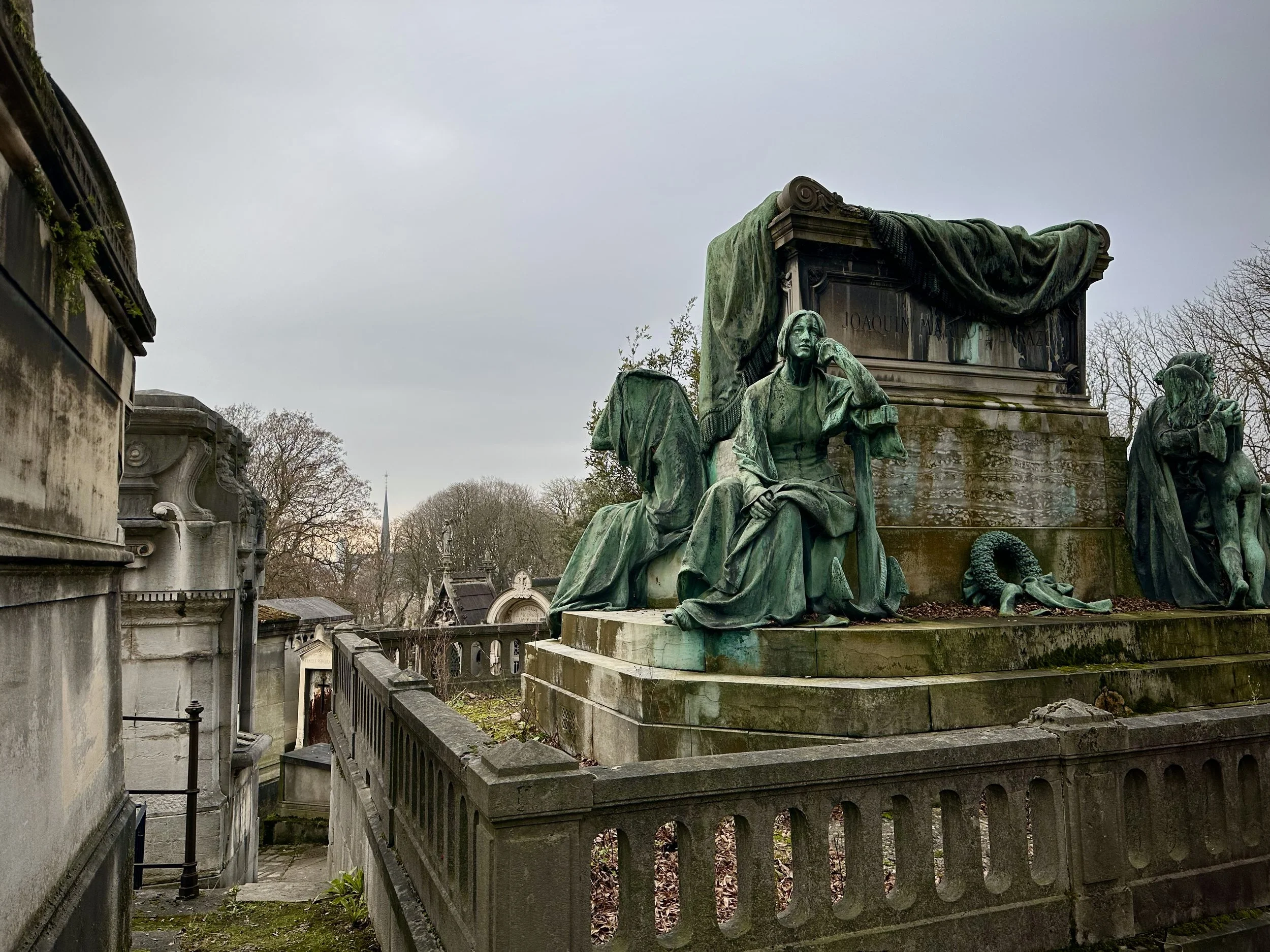 Historical monument with bronze statues, including a seated woman with a hand on her face, on a cloudy day in a cemetery or memorial park, with other tombstones and leafless trees in the background.