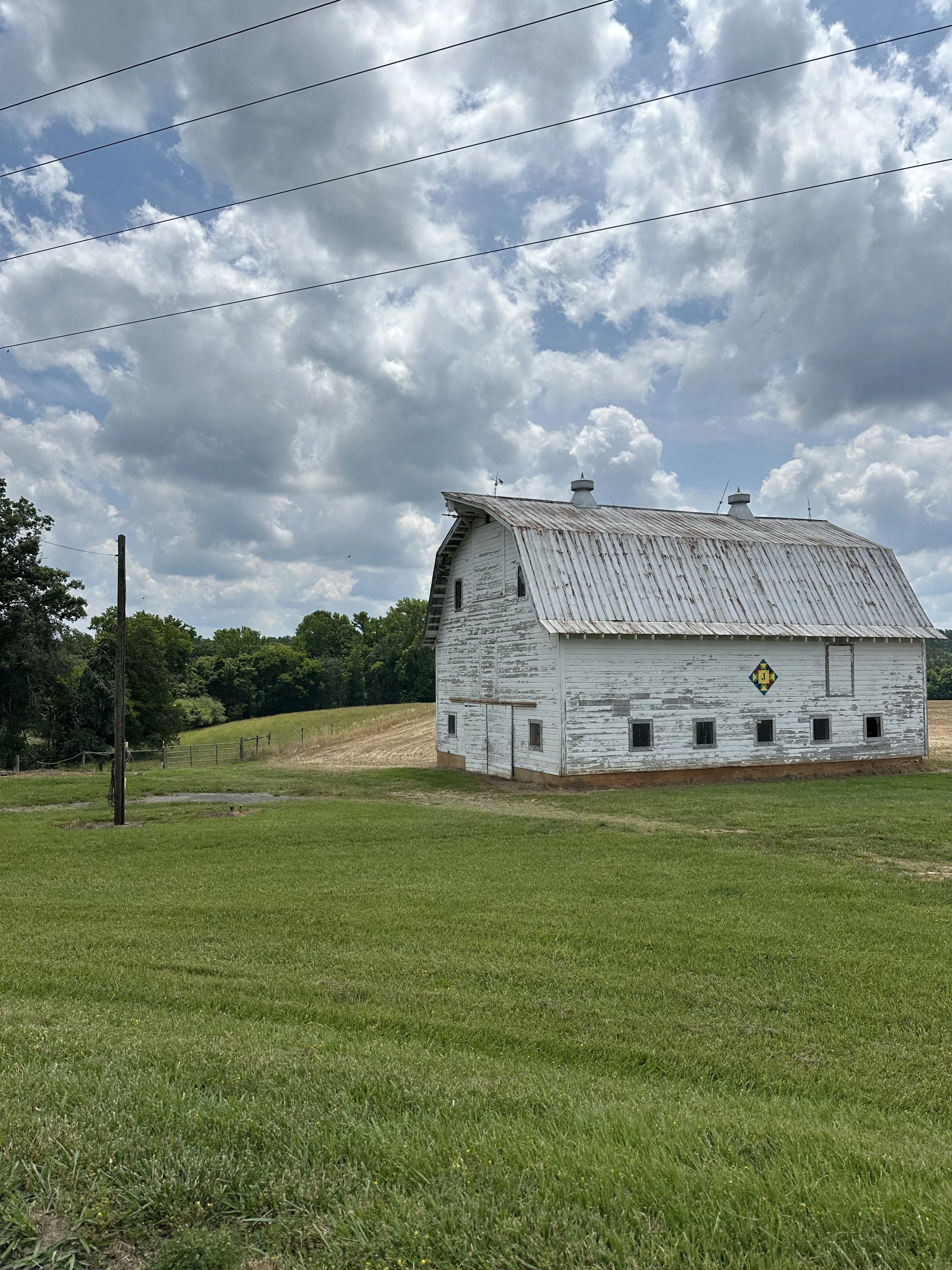 A white wooden barn with a rusty metal roof, situated on a grassy field under a partly cloudy sky with power lines overhead.