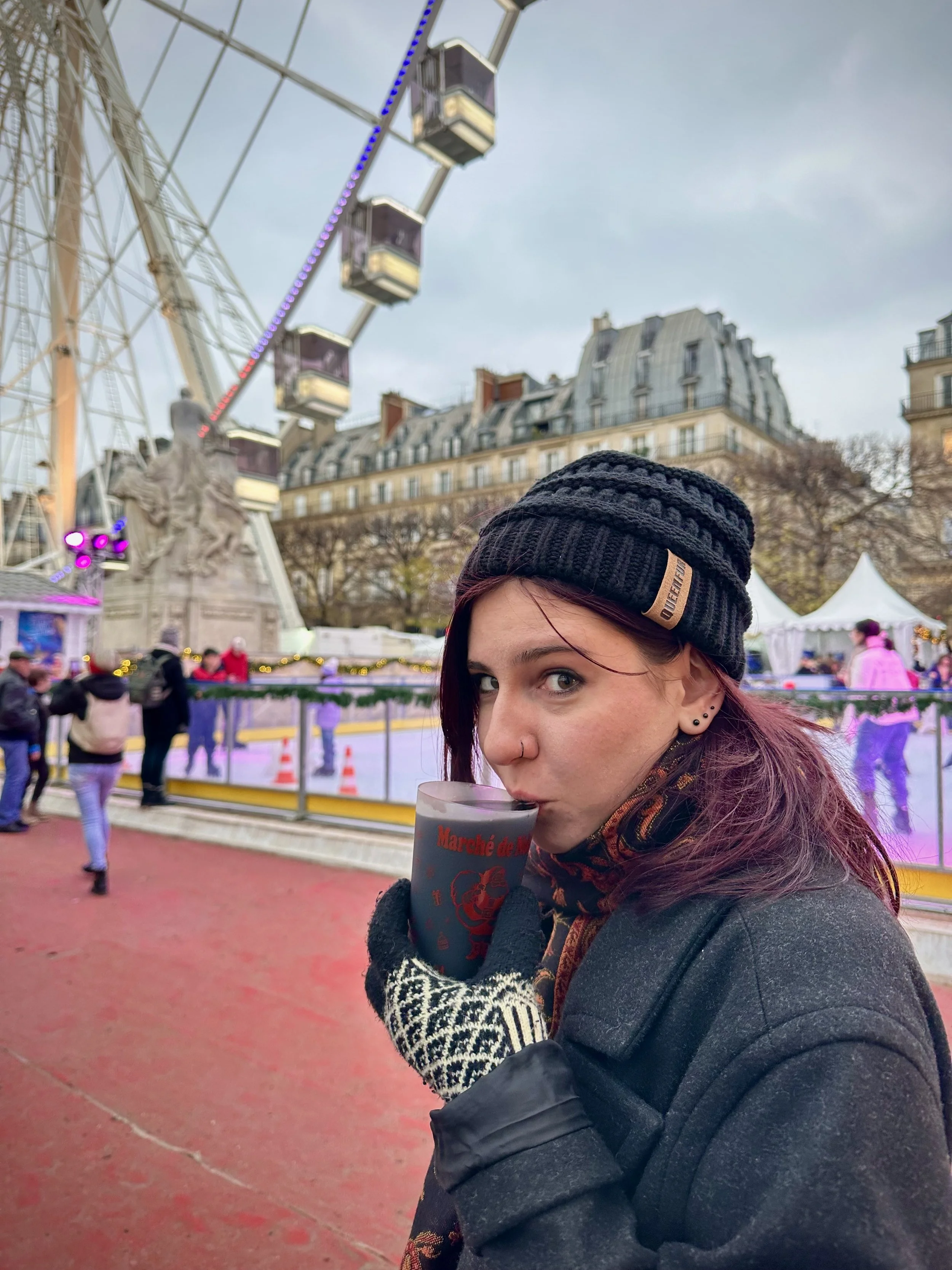 A woman with red hair, wearing a black beanie, black coat, and patterned gloves, drinks from a gray cup at an outdoor ice skating rink in front of a cityscape with a ferris wheel and historic buildings.