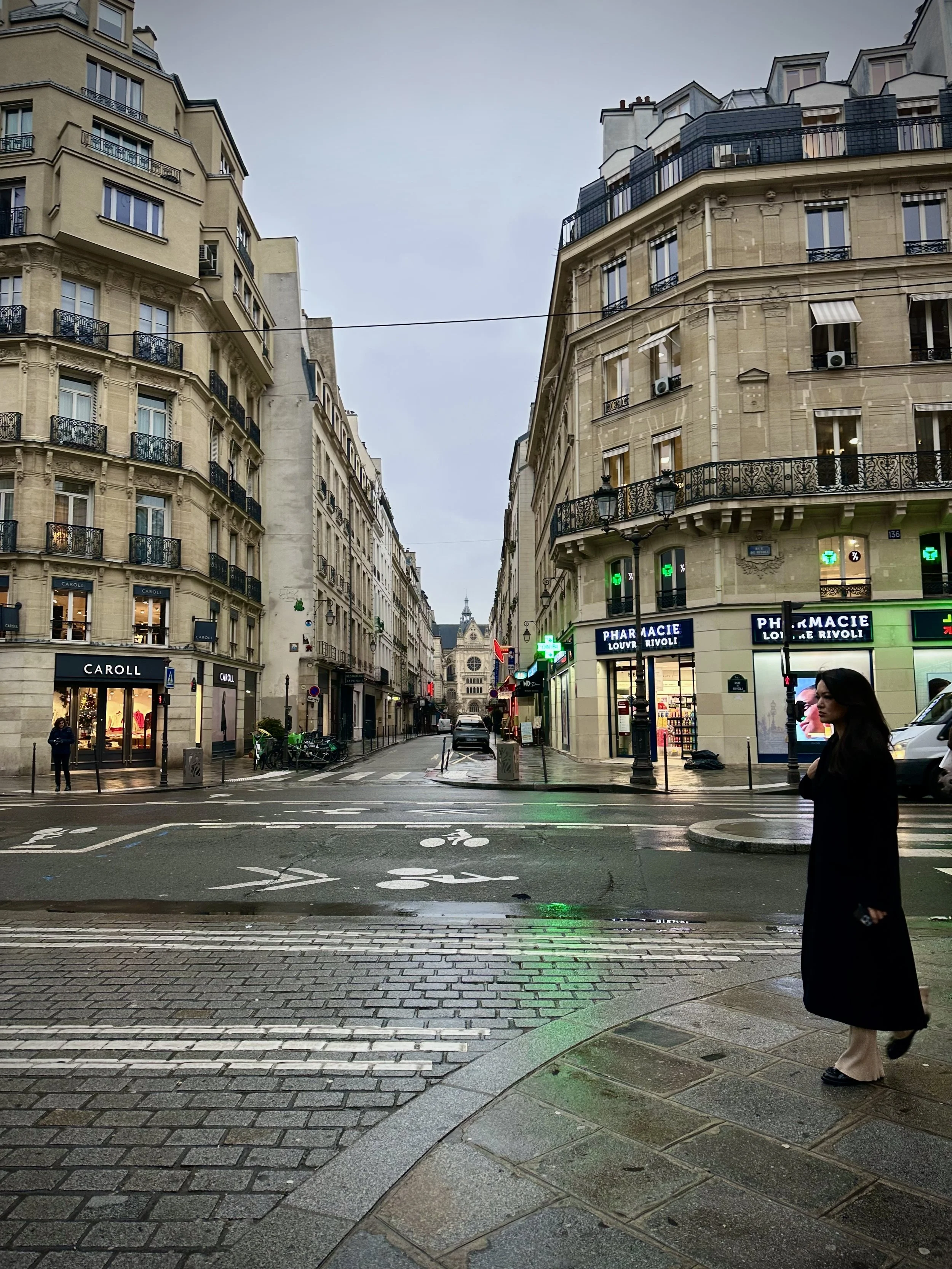 Paris street scene with Haussmann-style buildings, a woman in black coat crossing the street, and a pharmacy with green cross signs in the background.