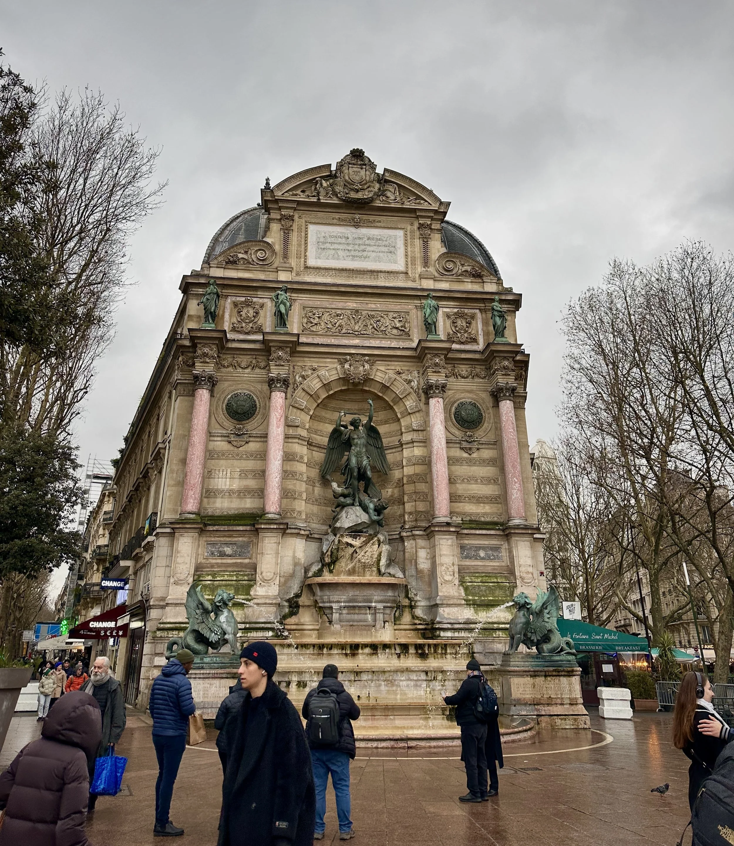 A historic building with ornate architecture and a large central fountain featuring a statue of a winged figure with water cascading from its base, surrounded by people walking on the street.