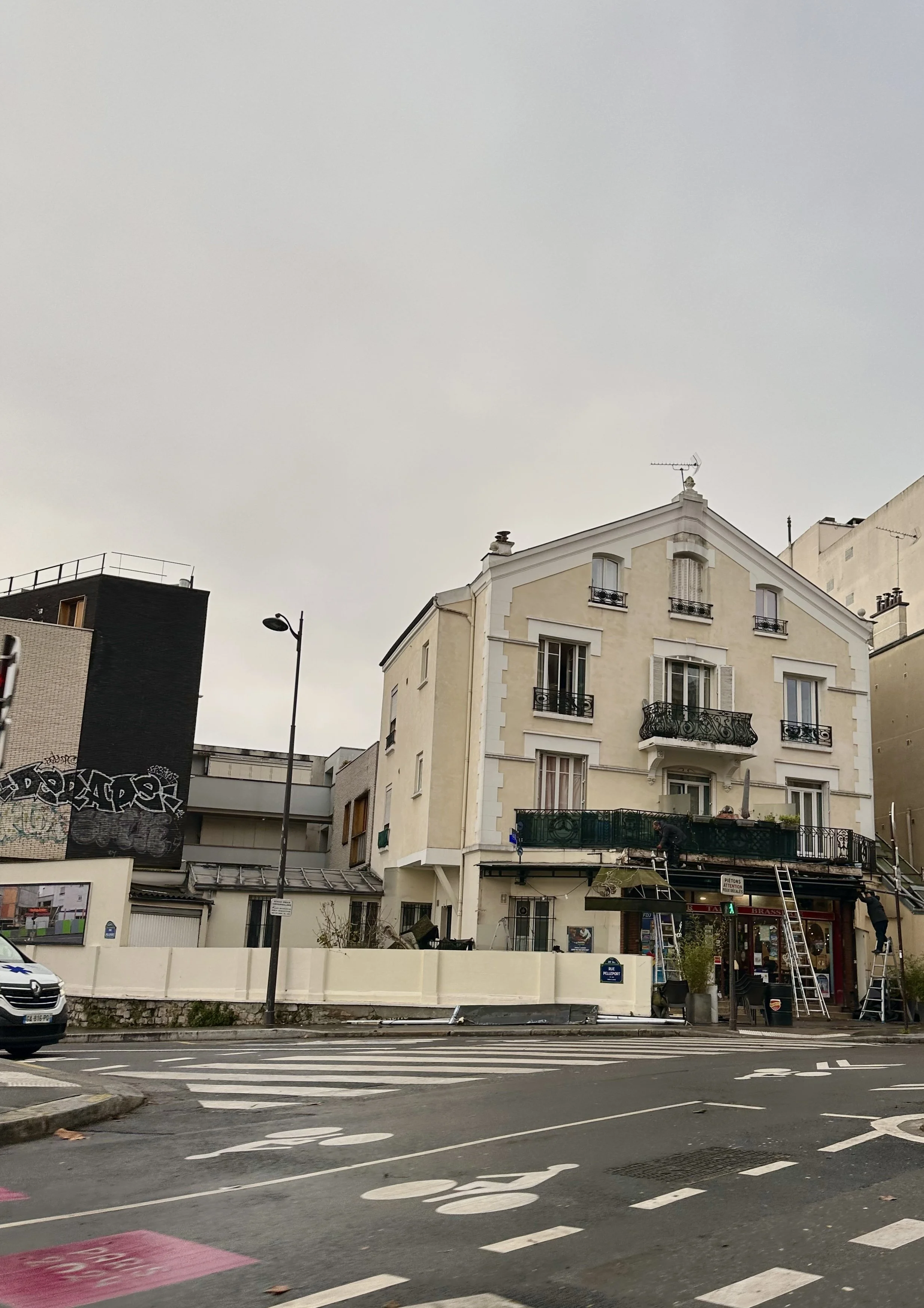 Multi-story beige building with awnings and balconies, situated at street corner with crosswalks and a parked white vehicle, overcast sky overhead.