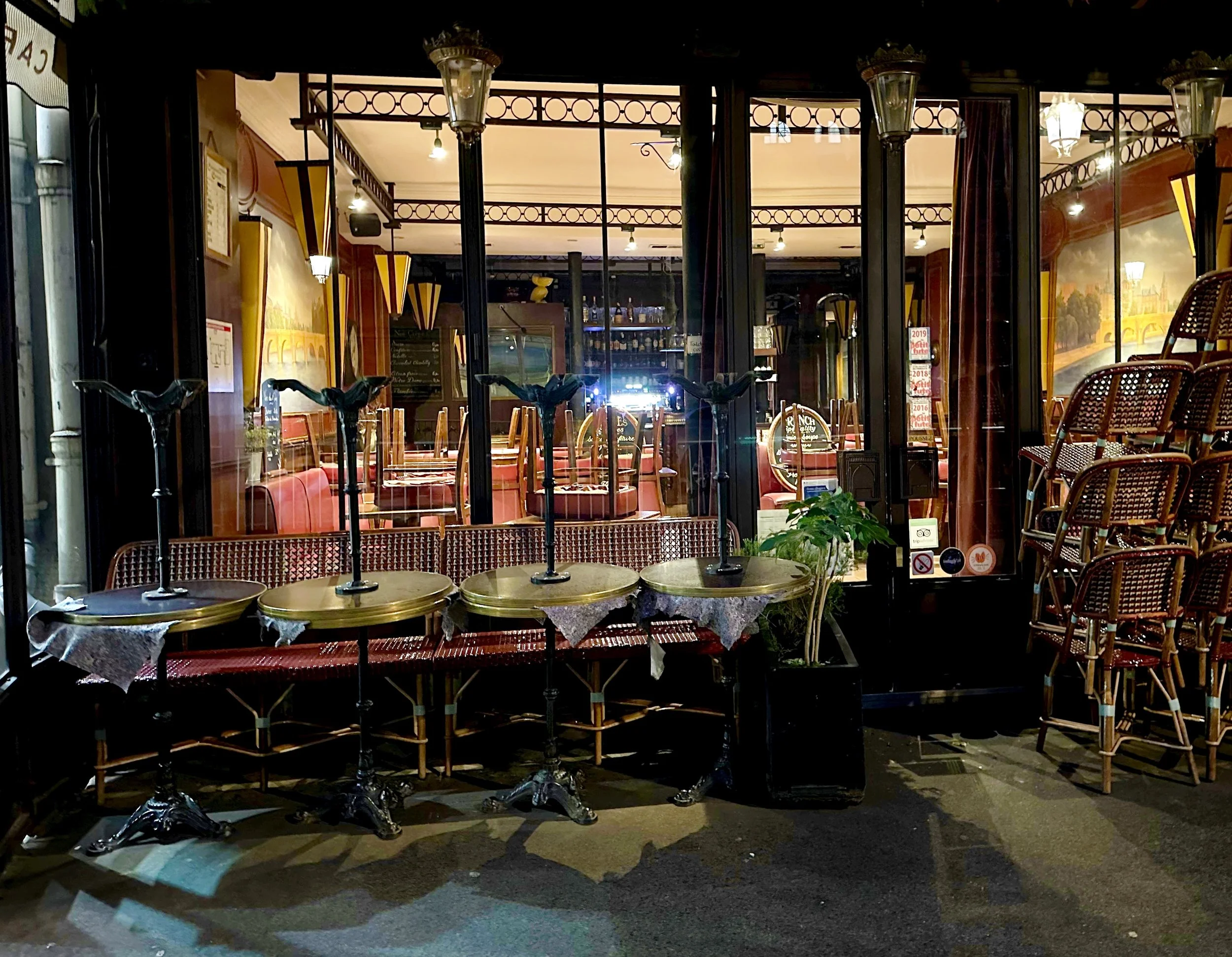 Outside view of a cozy café at night with tables and chairs stacked outside, illuminated by warm lighting, showing through large glass windows.
