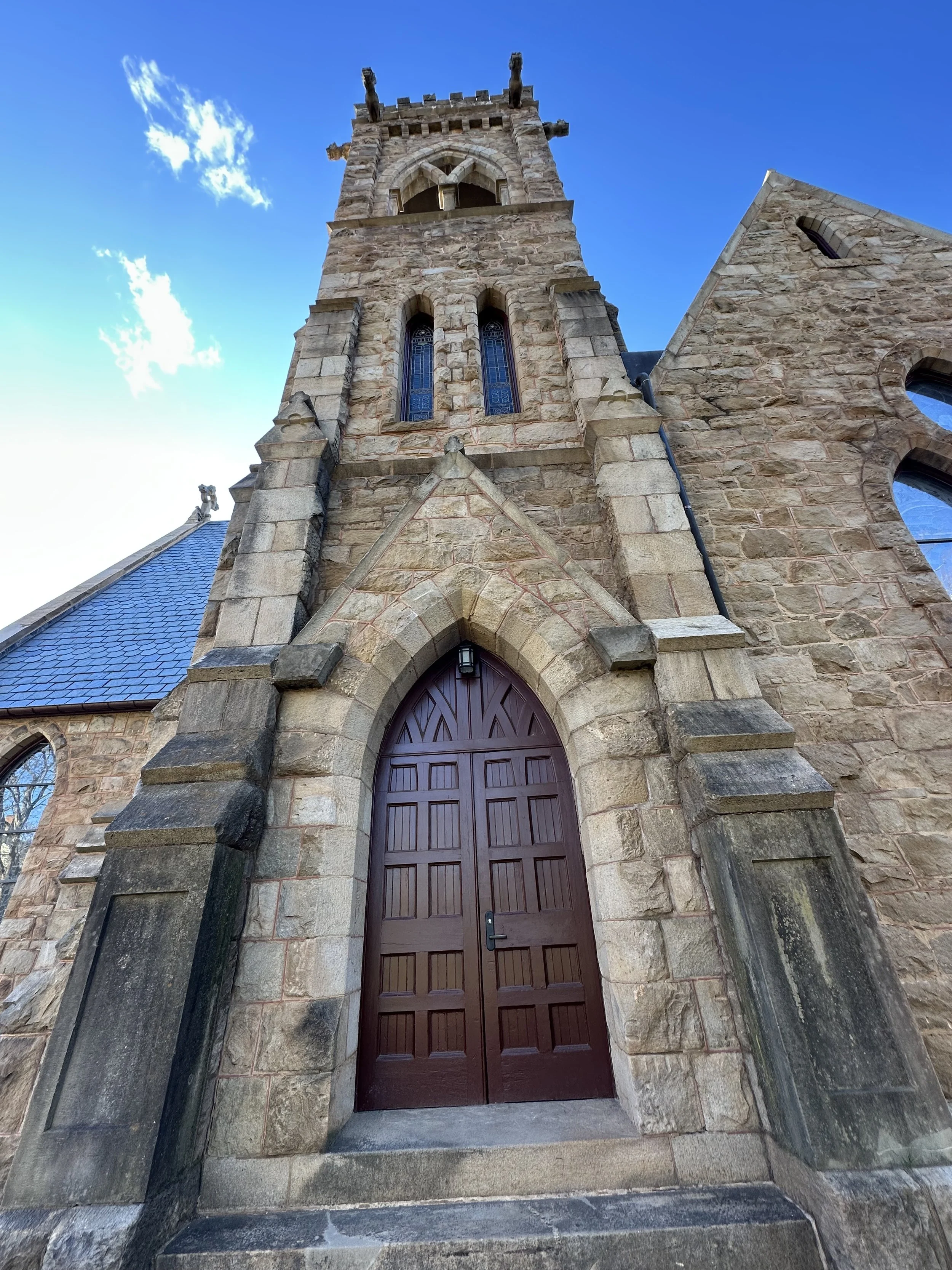 A low-angle view of UVA Chapel: wooden double door, tall stained glass windows, and a tall bell tower against a blue sky with a few clouds.
