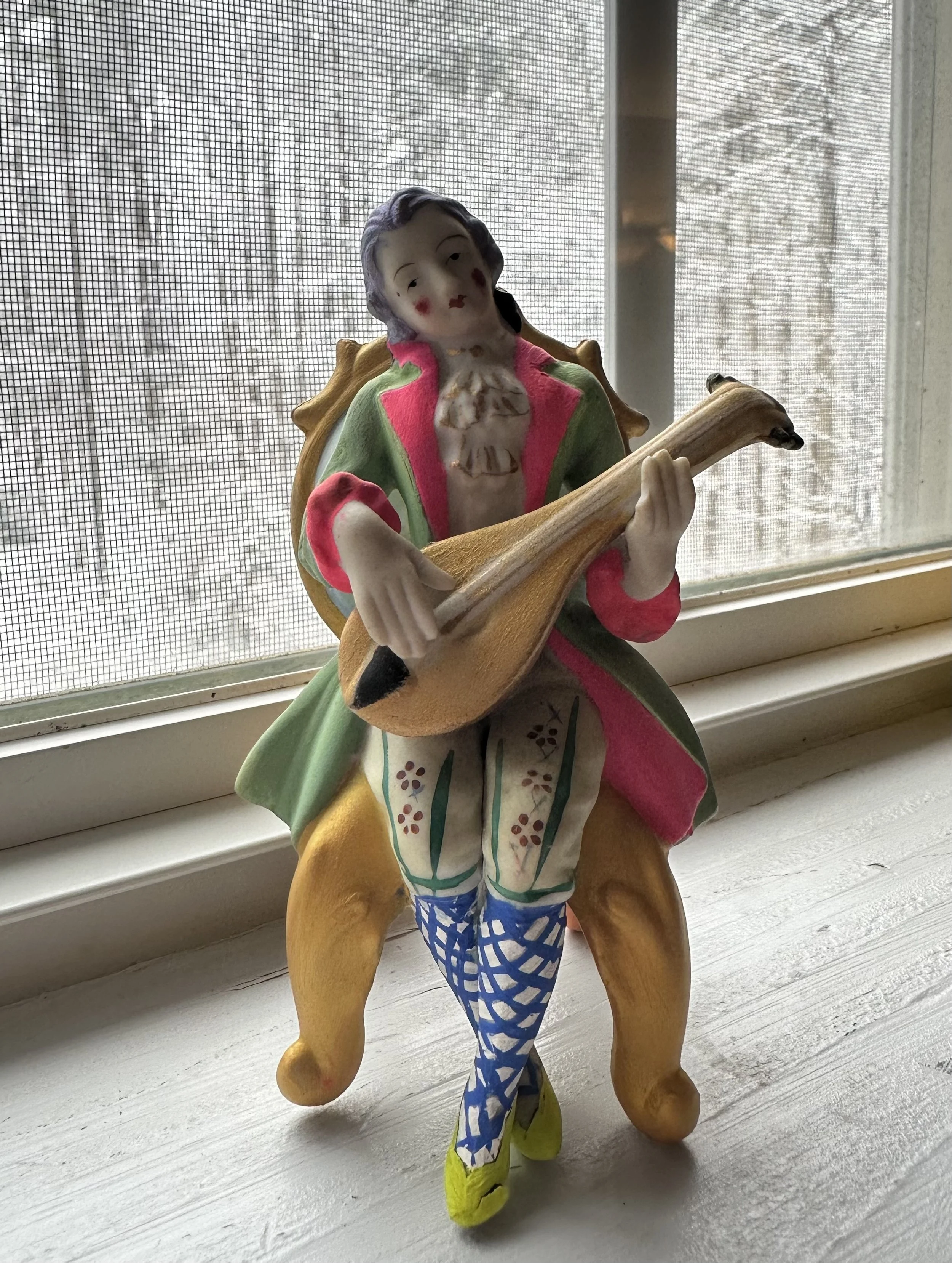 Colorful figurine of a man playing an acoustic guitar, dressed in vintage-style clothing with floral leggings and blue fishnet stockings, sitting on a wooden surface near a window.