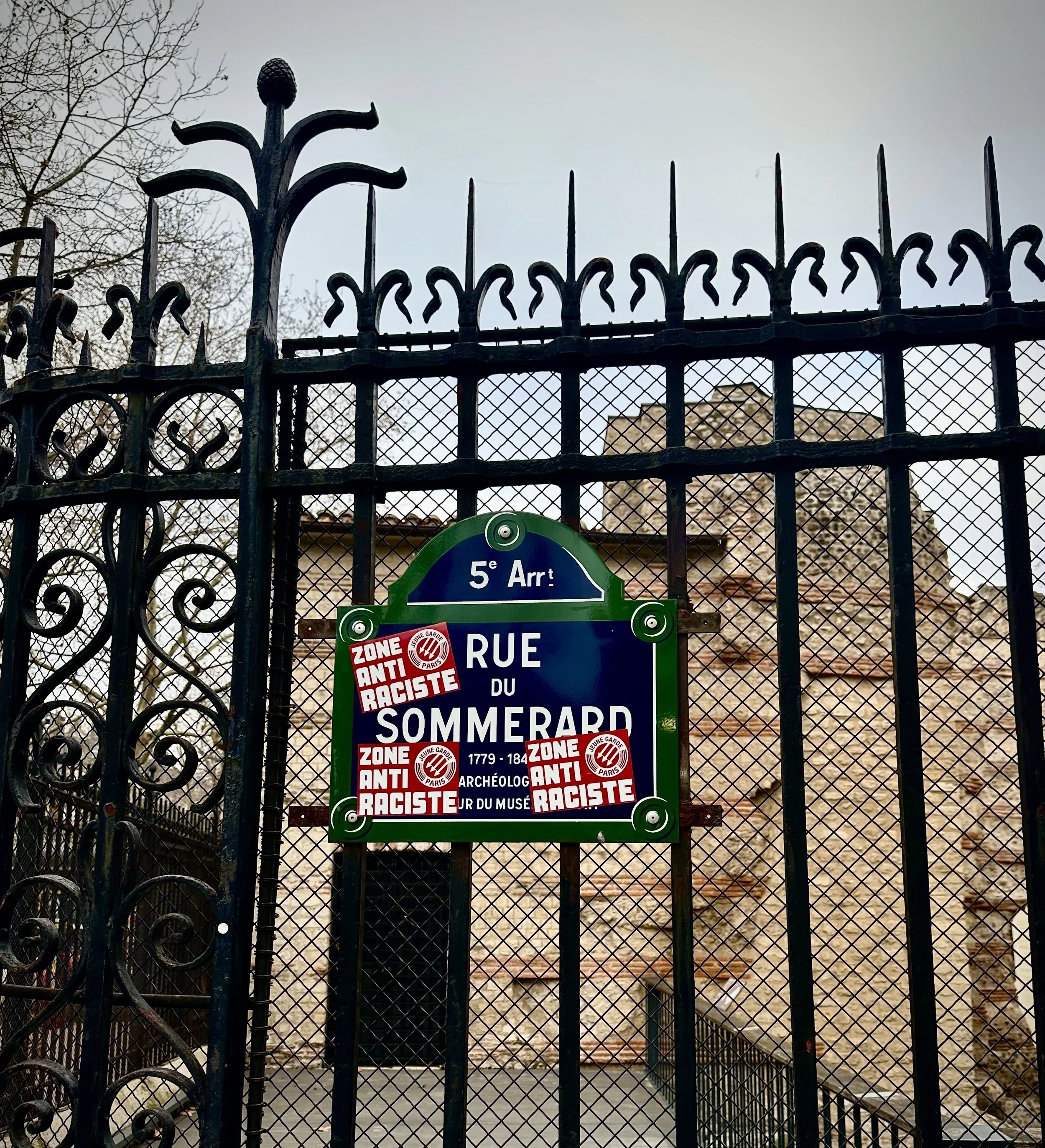 Street sign for Rue du Sommerard on a black iron fence, with three stickers reading 'Zone Anti Raciste'. In the background, a stone building with arches and leafless trees.