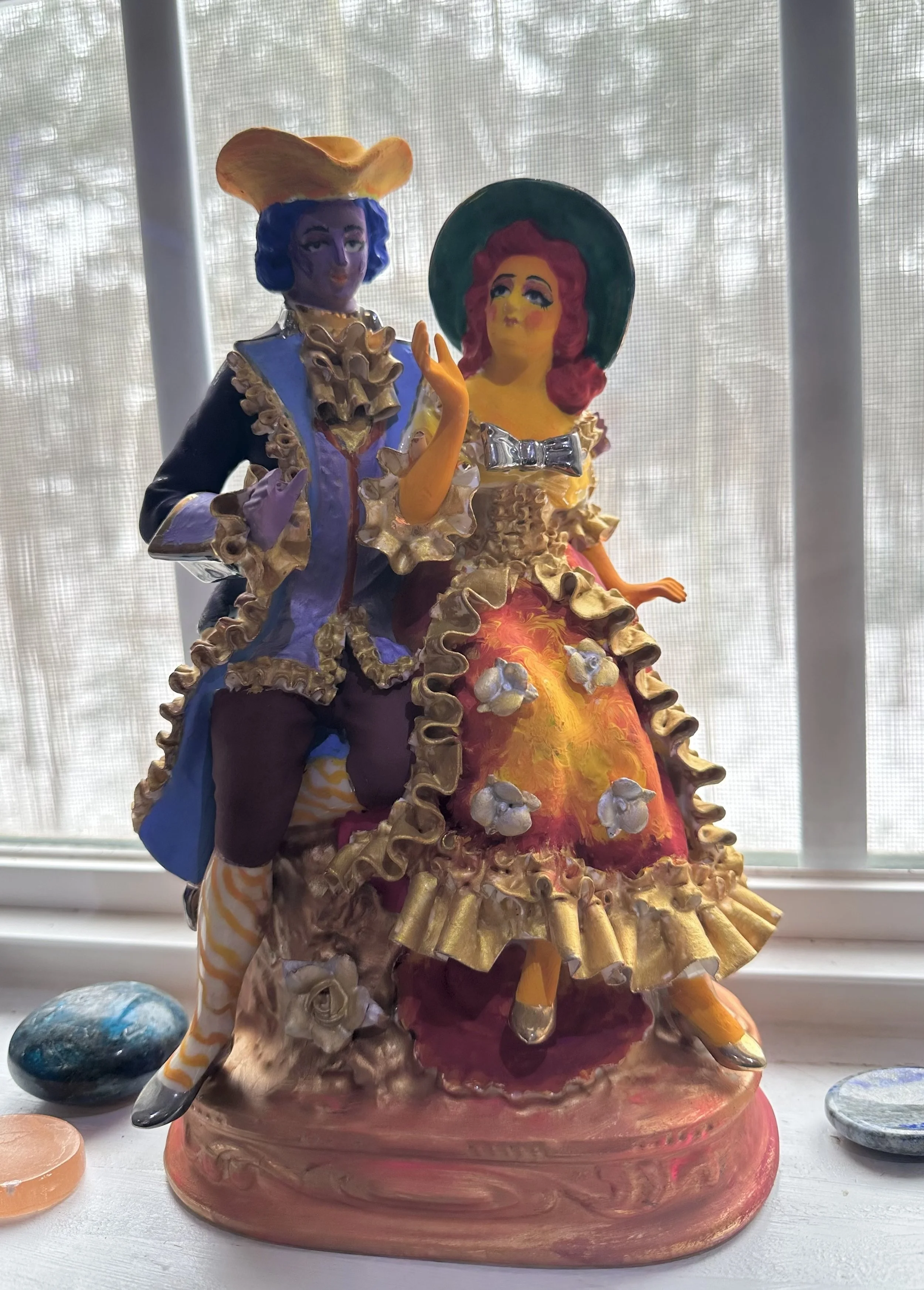 Colorful ceramic sculpture of a man and a woman dressed in elaborate 18th-century style costumes, standing on a decorative base, with a window and trees in the background.
