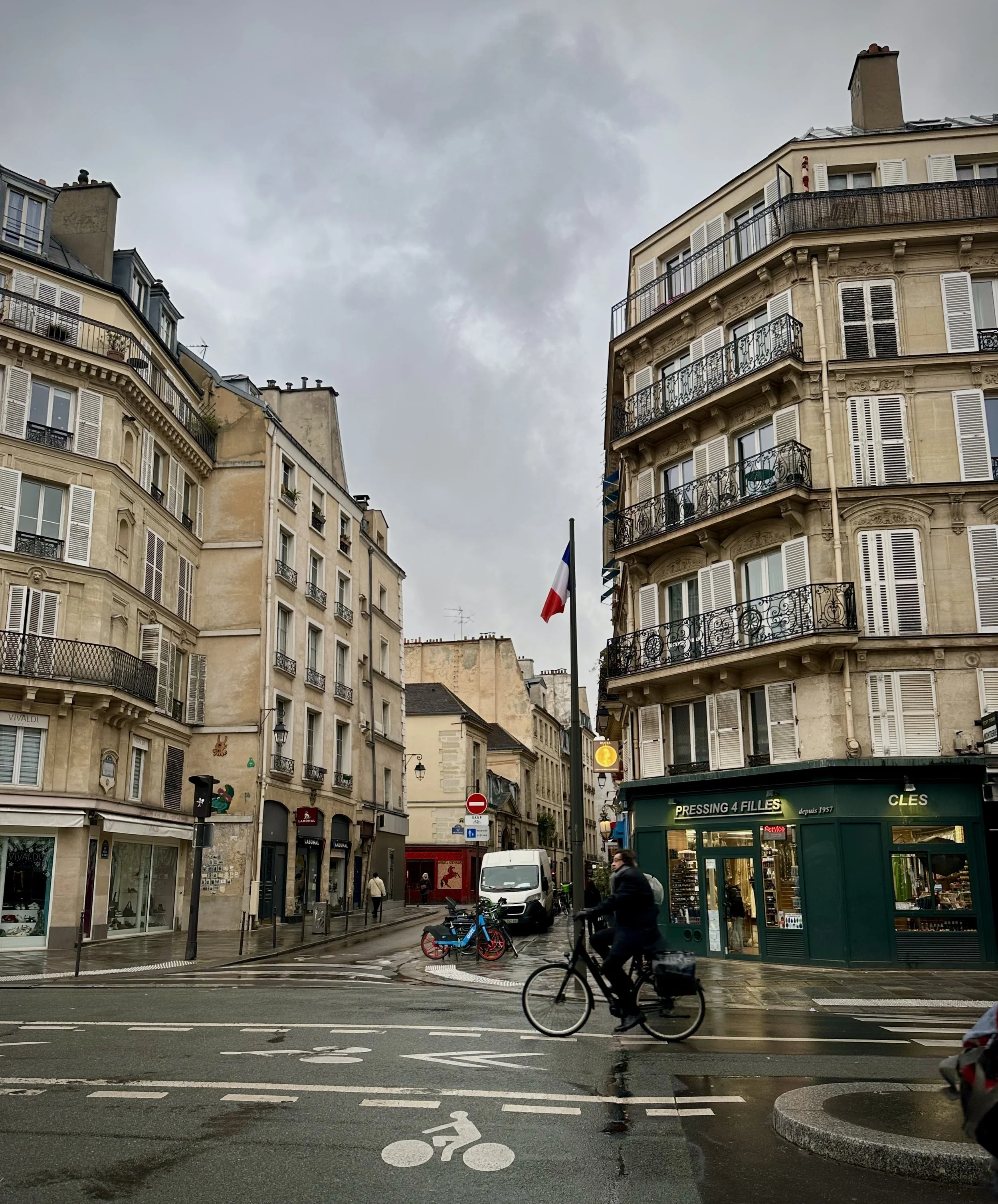 Overcast street scene in Paris with classic apartment buildings, a French flag, a person riding a bicycle, and storefronts.